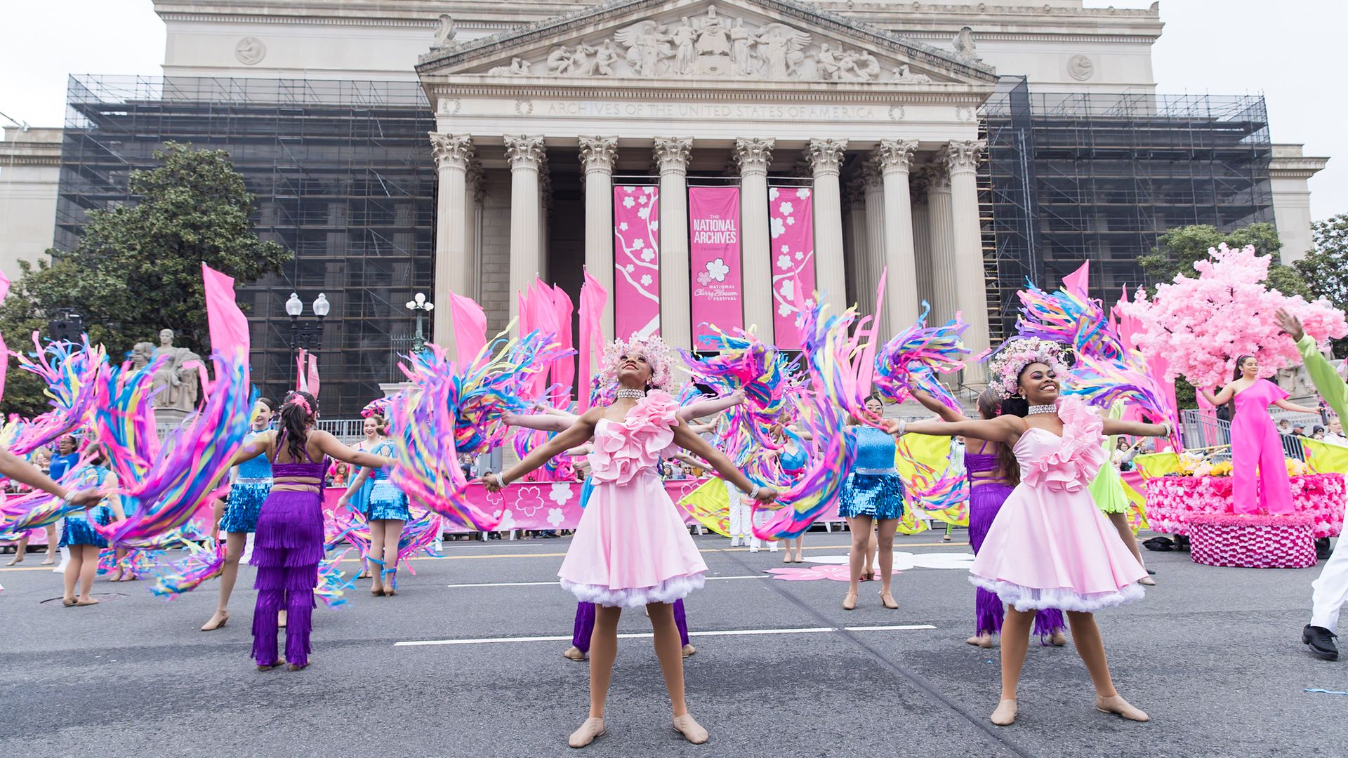 Parade scene in front of a grand neoclassical building with tall columns. Dancers in pink and purple dresses wave multicolored streamers; pink banners hang between the pillars.