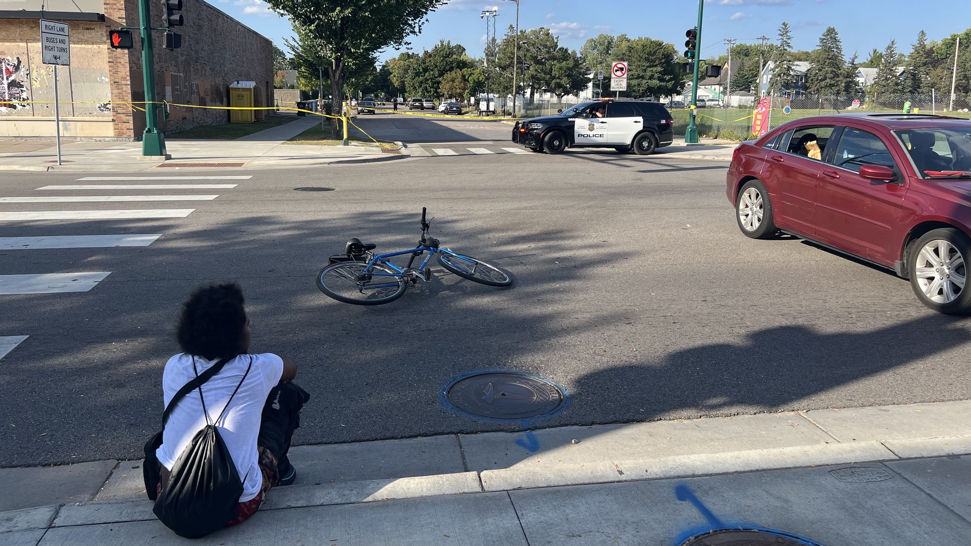 A person sits on a sidewalk near a fallen blue bicycle in the middle of an intersection, where police tape blocks off the area and a black-and-white police SUV is parked nearby on a sunny day.