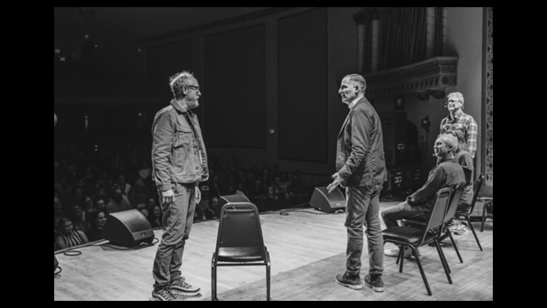 Black-and-white photo of four men on a theater stage: two stand facing each other center stage, one sits on a chair to the right, audience seated in the dim background, ornate stage side.