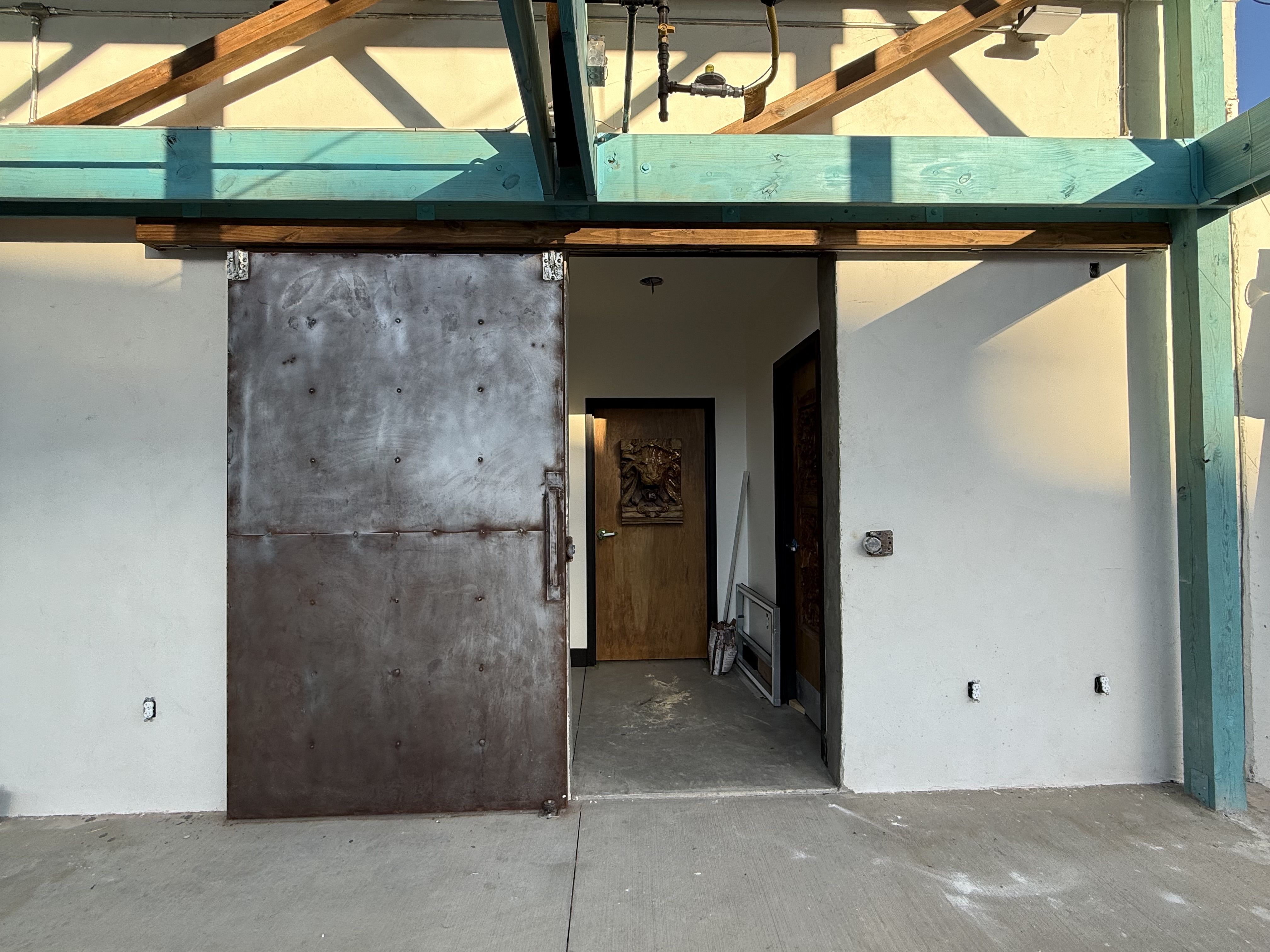 Industrial entrance with a large rusty metal sliding door partially open, revealing a small hallway with a wooden door featuring carved artwork inside. Turquoise wooden beams overhead.