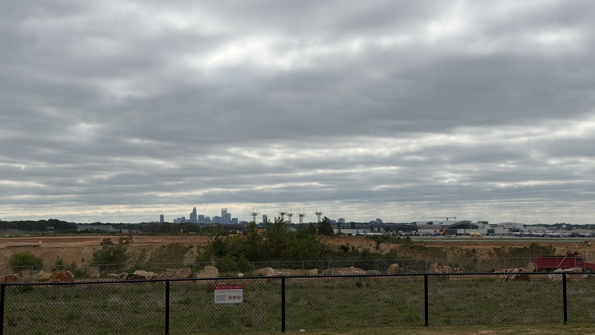 Overcast sky with thick gray clouds over a distant city skyline; a chain-link fence and grassy, rocky foreground sit before an airport with aircraft and terminal buildings on the horizon.
