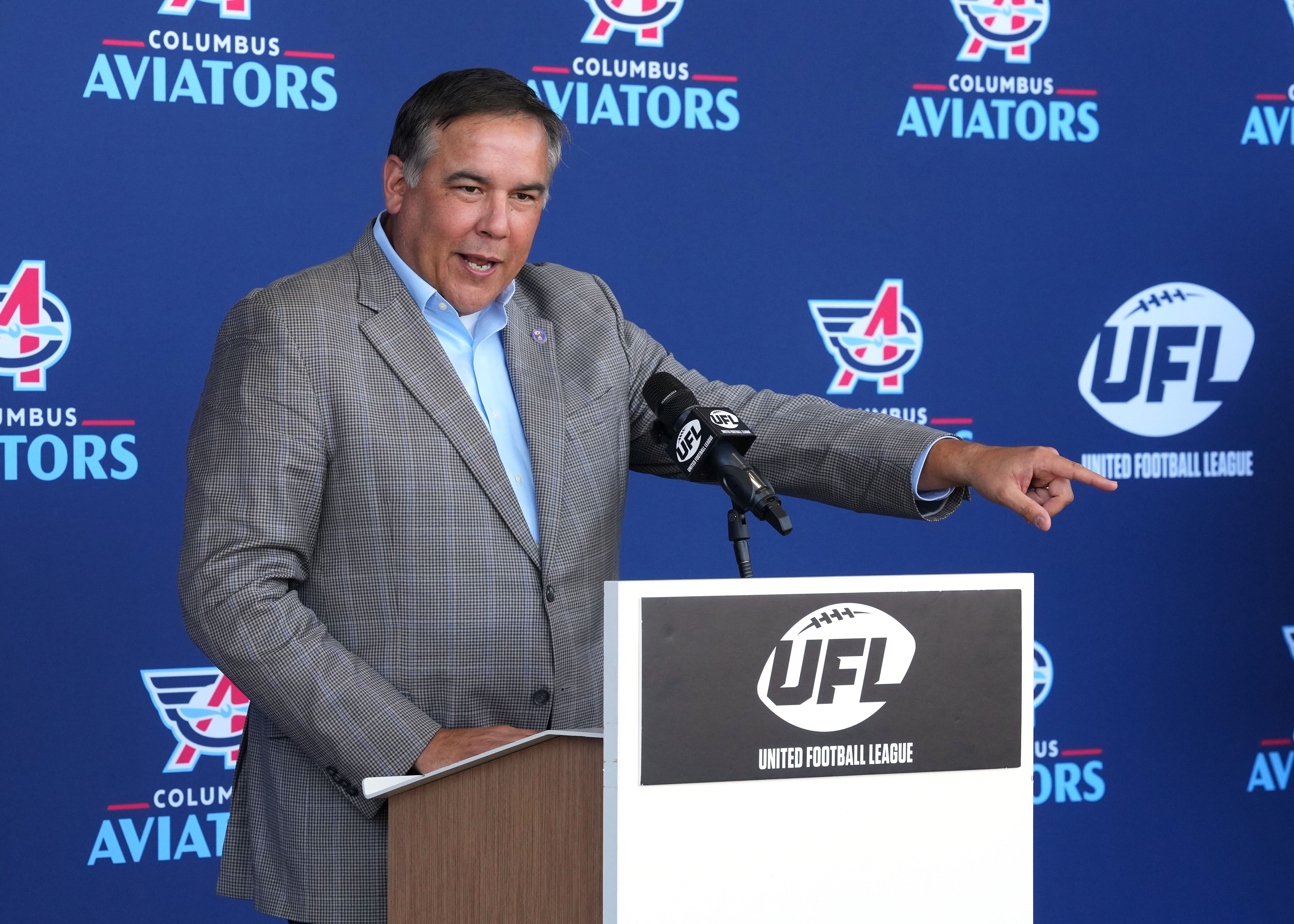 Columbus Mayor Andrew Ginther speaking at a podium with a "UFL United Football League" sign, pointing to the right. Blue backdrop with Columbus Aviators logos.
