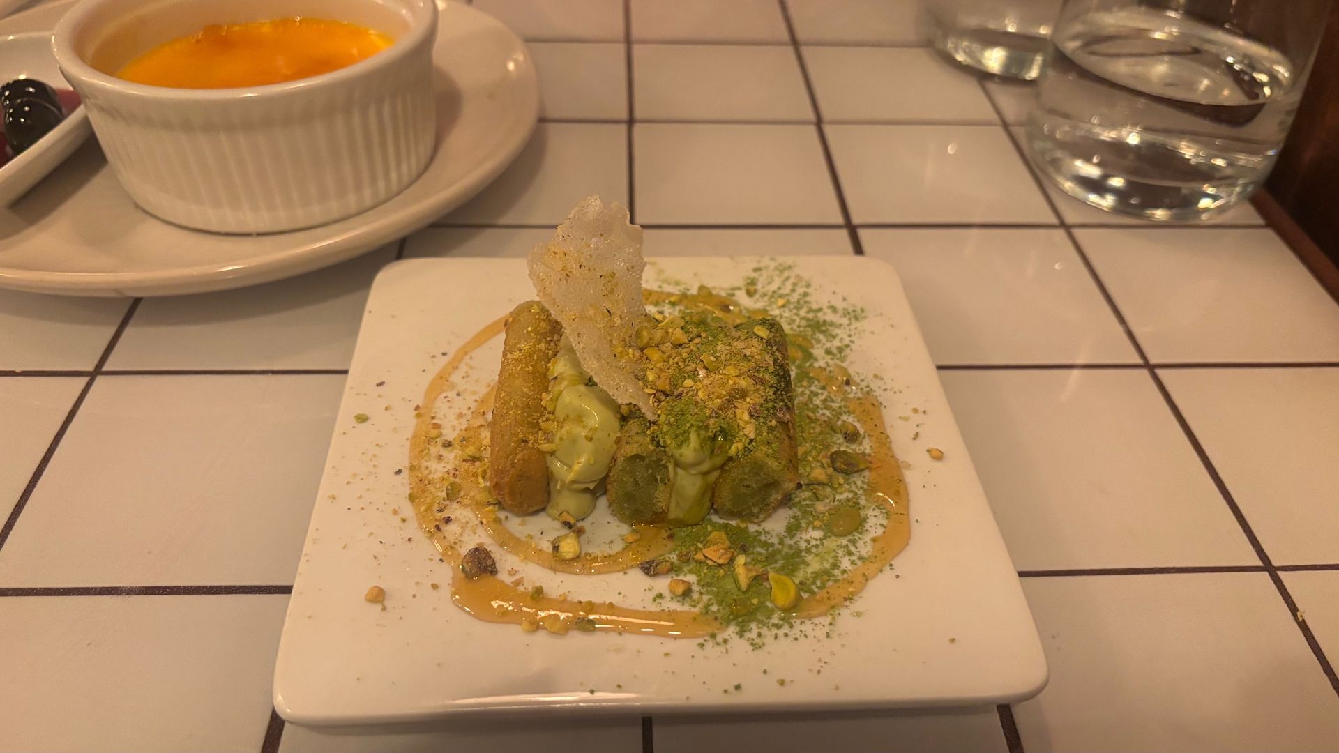 A plated dessert on a white square dish: layered pastry topped with pistachio dust, a crisp sugar shard, and golden caramel sauce; in the background a ramekin of orange custard and a glass of water.