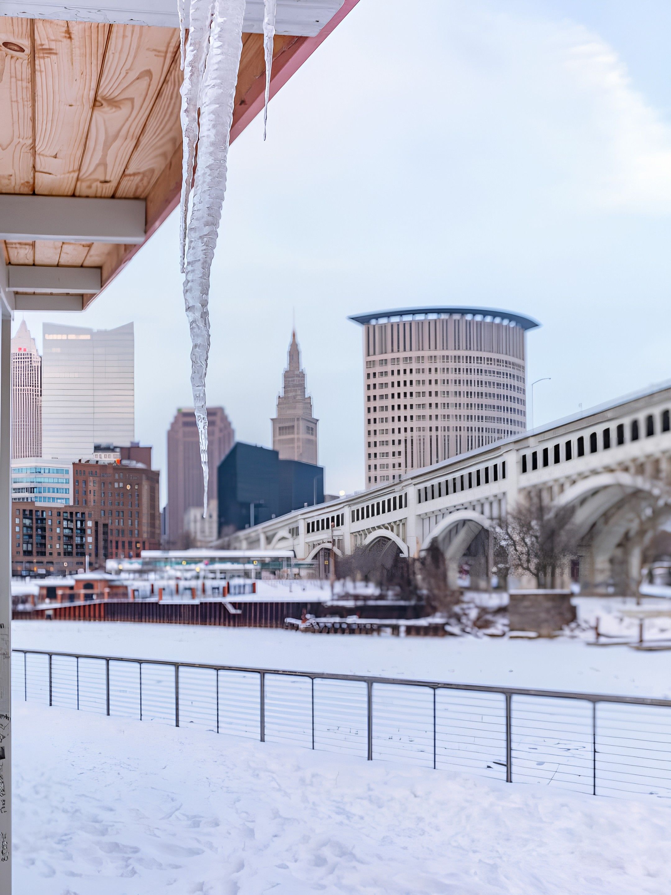 Snow-covered riverside with large icicles hanging from a wooden roof, a white arched bridge, and city buildings in the background under a pale blue sky.
