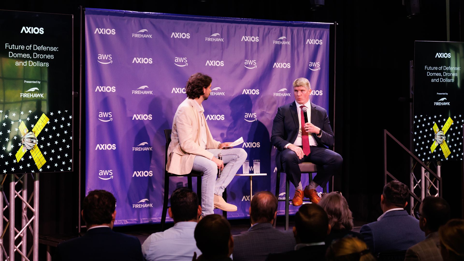 Two men in a discussion on stage at an Axios event titled Future of Defense: Domes, Drones and Dollars, with logos of Axios, Firehawk, and AWS on a purple backdrop and screens.