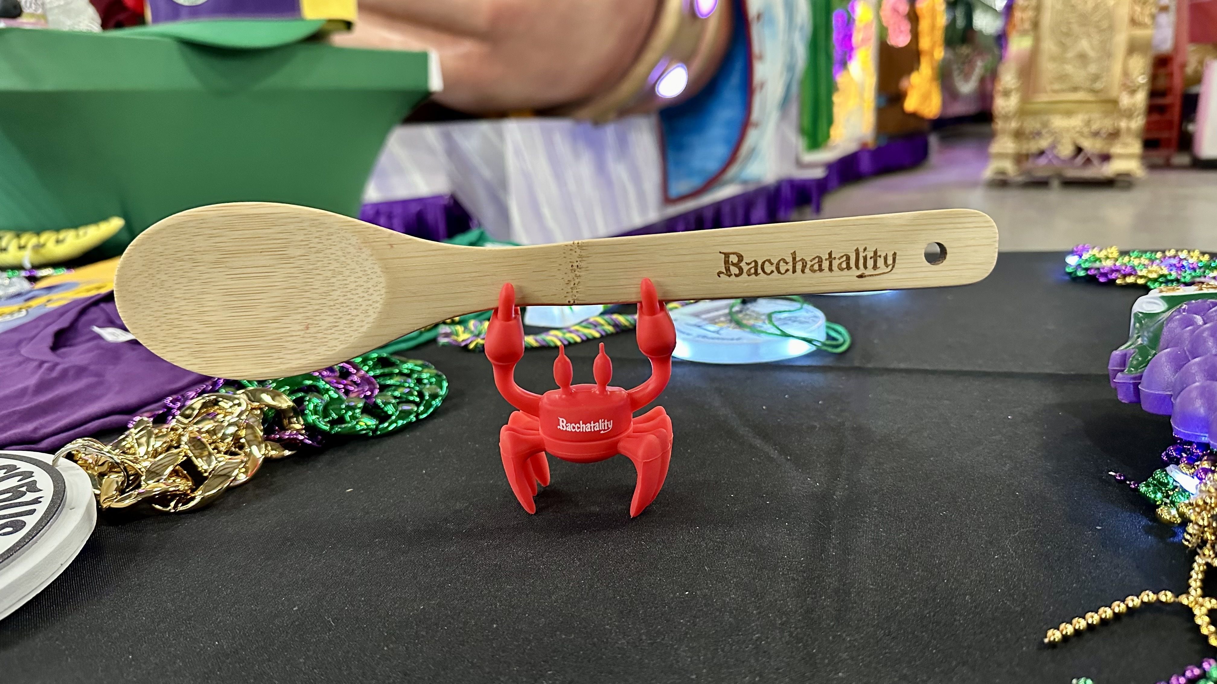 A small red crab holds up a wooden spoon that says "Bacchatality."