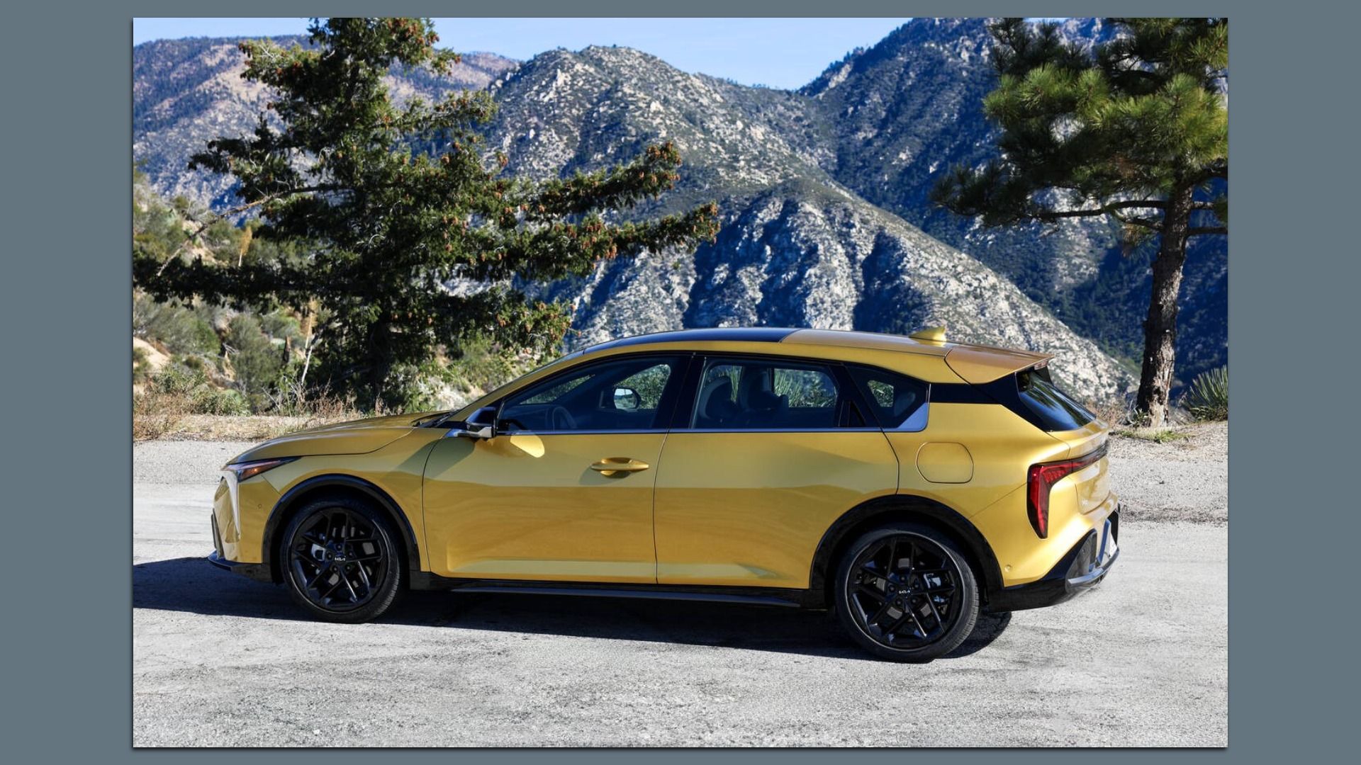Side view of a golden yellow Kia K4 hatchback with black wheels parked on a gravel road, with green pine trees and rocky mountains in the background under a clear blue sky.