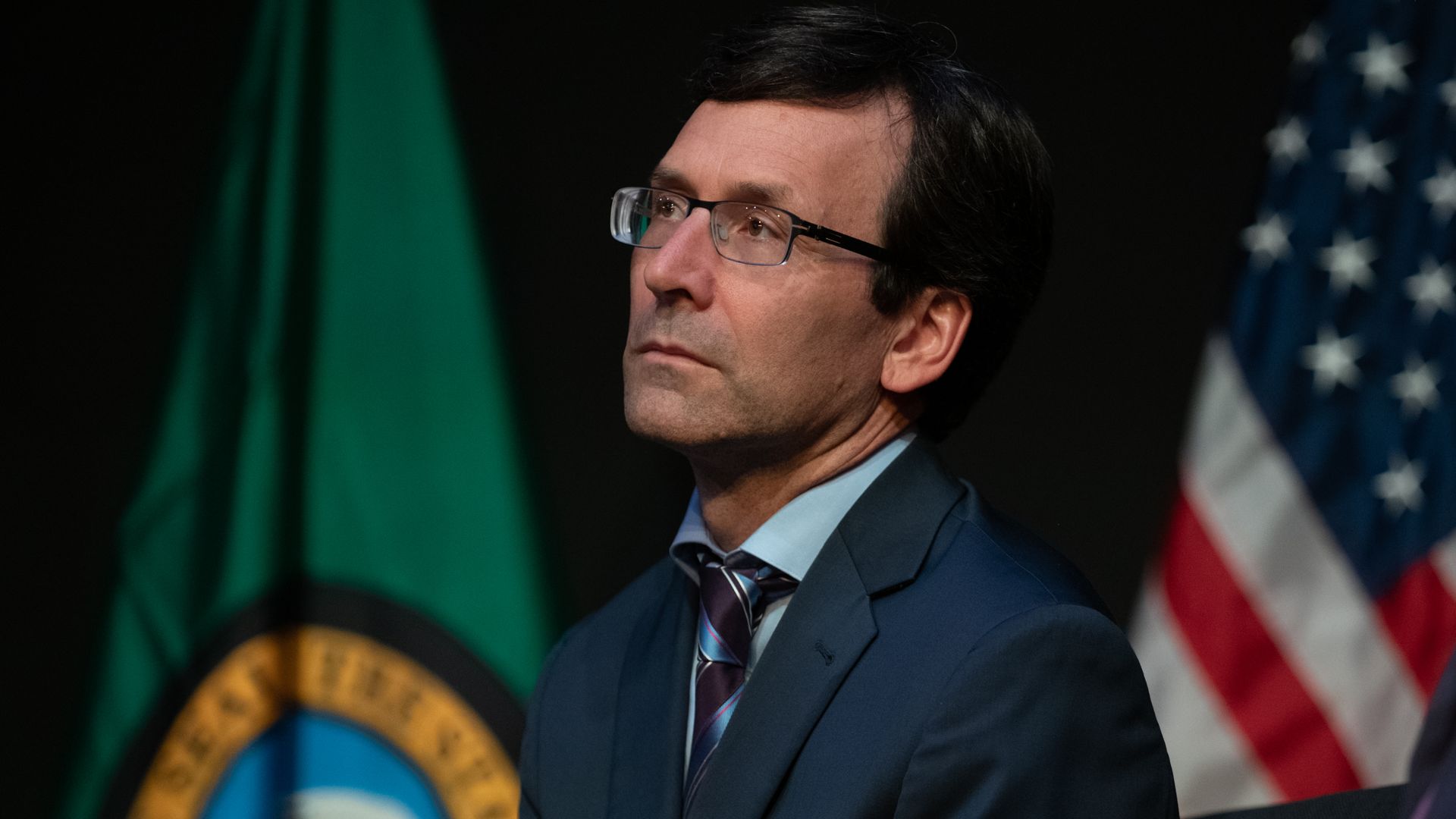 Bob Ferguson, shown with glasses in a dark blue suit and striped tie, sits thoughtfully with Washington state and American flags blurred in the background.