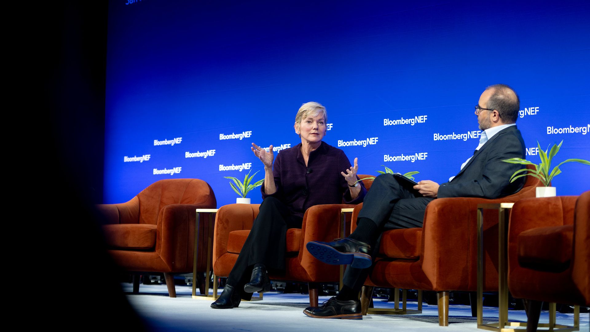 Two people seated on orange chairs on a stage at the BNEF Summit in San Francisco with a blue background displaying BloombergNEF logos, engaged in a discussion.