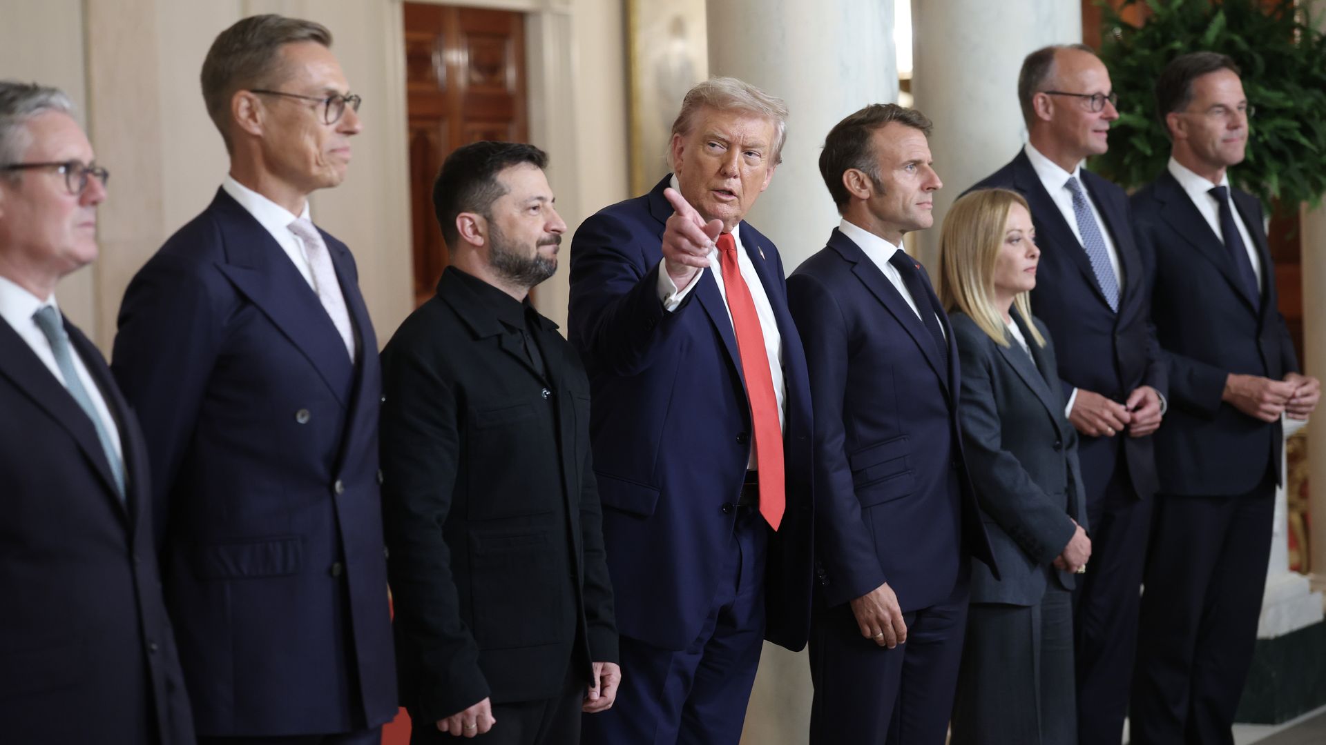 A group of eight formally dressed politicians and leaders stand indoors. One man in the center wearing a navy suit and red tie is pointing. The others wear dark suits or jackets.
