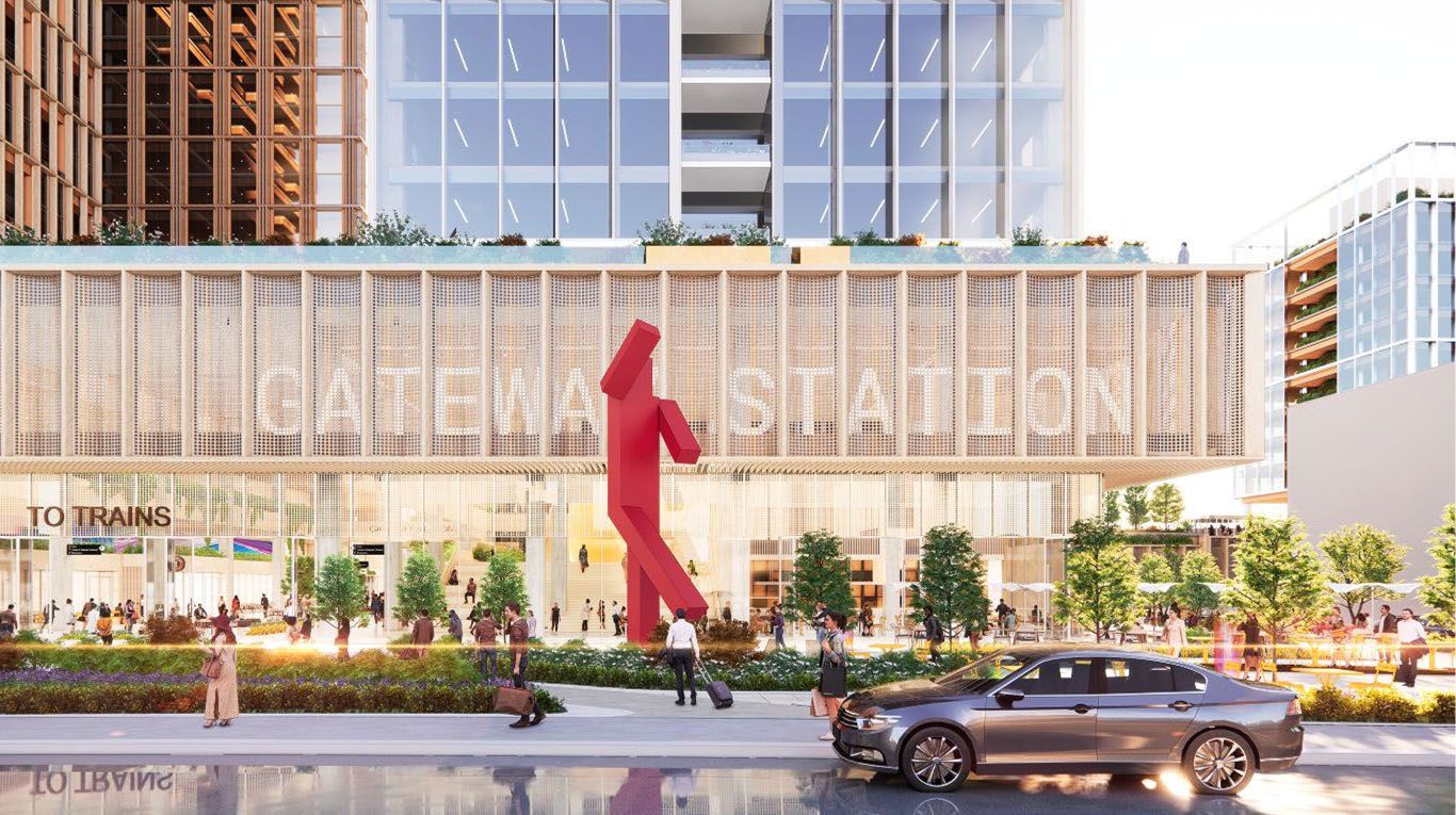 Modern station entrance with large glass windows and a vertical wooden grid reading "GATEWAY STATION." People walk near green bushes and trees, a red abstract sculpture, and a parked gray car.