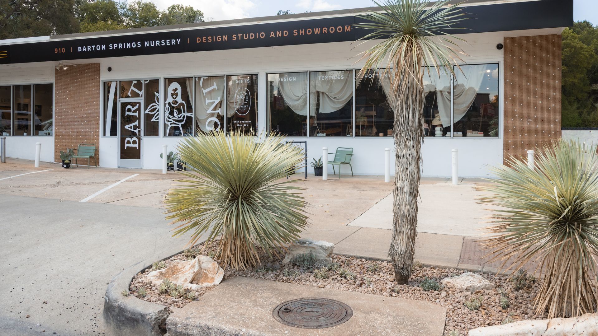 Single-story showroom building with a long black sign that reads Barton Springs Nursery | Design Studio and Showroom. Large windows with white drapes, a tall palm tree, and desert landscaping in front.