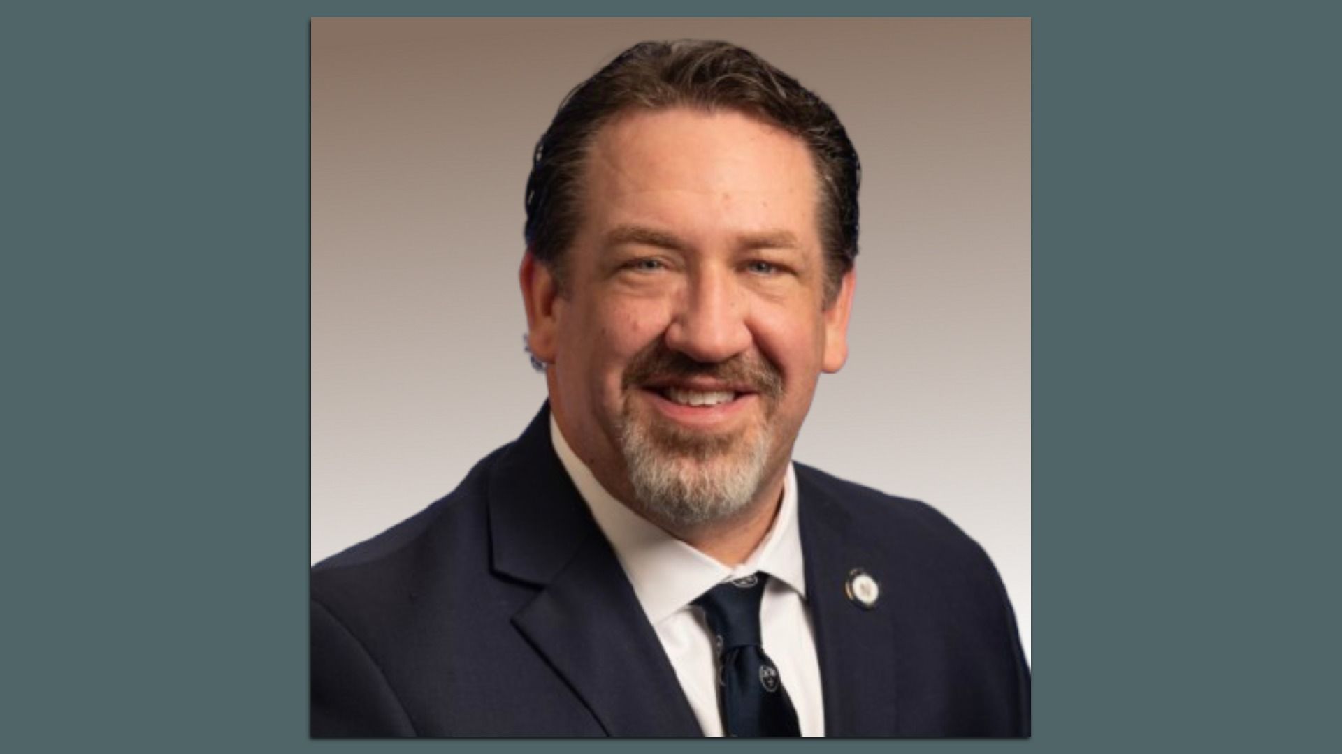 Portrait of a smiling man with short dark hair and a gray goatee, wearing a dark suit, white shirt, and navy tie, with a lapel pin, against a beige gradient background.