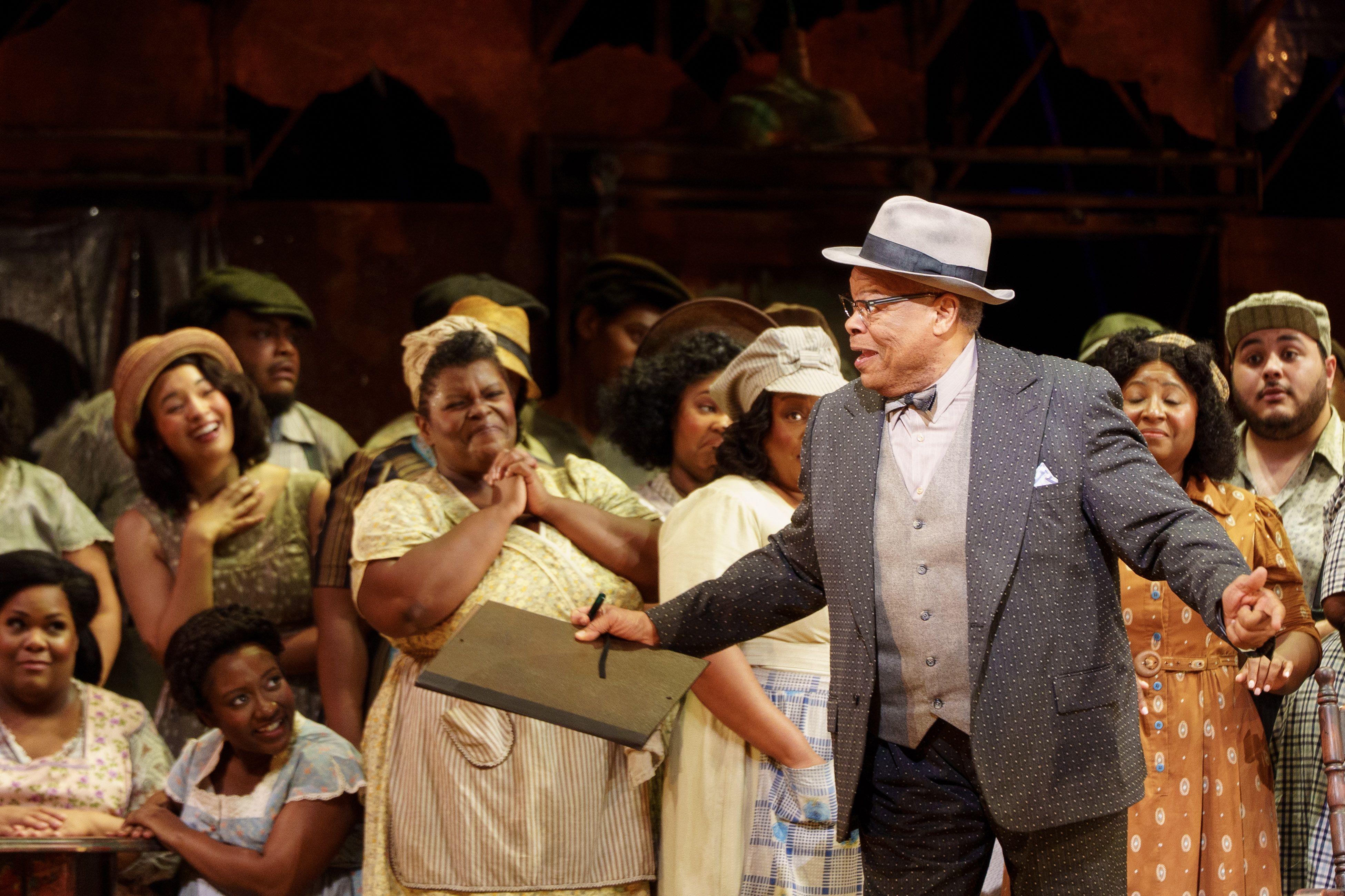A man in a gray polka-dot suit and hat speaks animatedly to a group of smiling and engaged people dressed in vintage clothing, suggesting a lively theatrical performance.