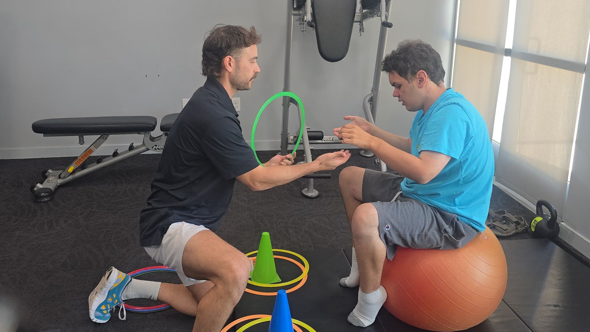 A man in a black shirt and white shorts kneels holding a green ring as another man in a blue shirt and gray shorts sits on an orange exercise ball in a gym with workout equipment and cones.
