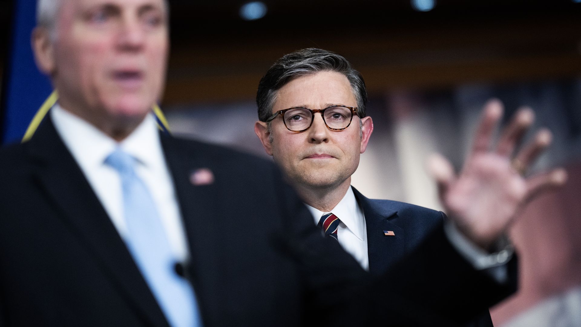 House Speaker Mike Johnson looking over the shoulder of Majority Leader Steve Scalise, wearing a blue suit and standing in front of a mural.