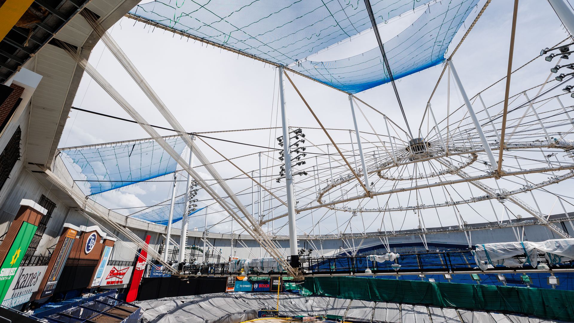 View of a baseball stadium under construction with the skeleton of a former roof above, featuring white beams and blue netting over the field area, empty seating, and advertising signs visible.