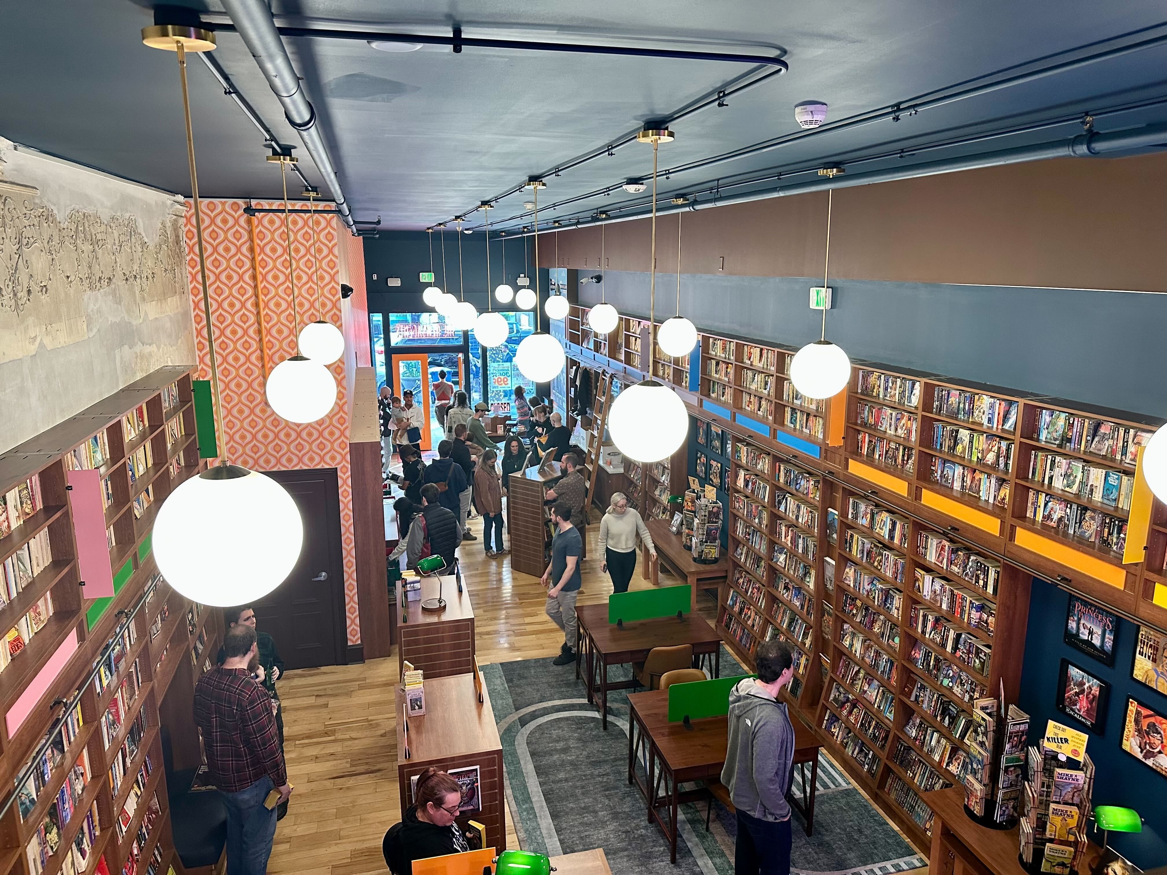 Interior of a busy bookstore with wooden shelves filled with books, round white hanging lights, orange patterned wall, tables with green dividers, and people browsing and talking.