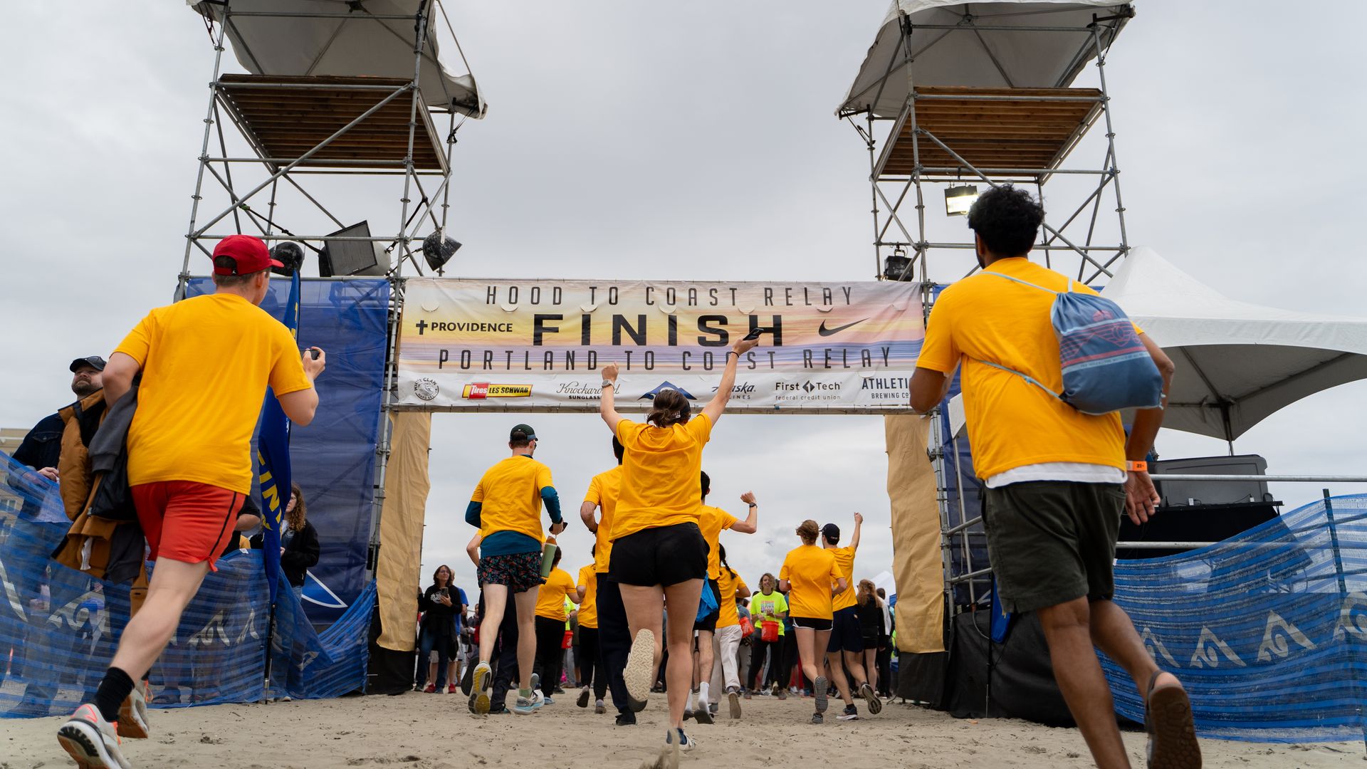 Runners wearing yellow shirts cross the finish line at the Hood to Coast Relay, Portland to Coast Relay event under a cloudy sky on a sandy path.