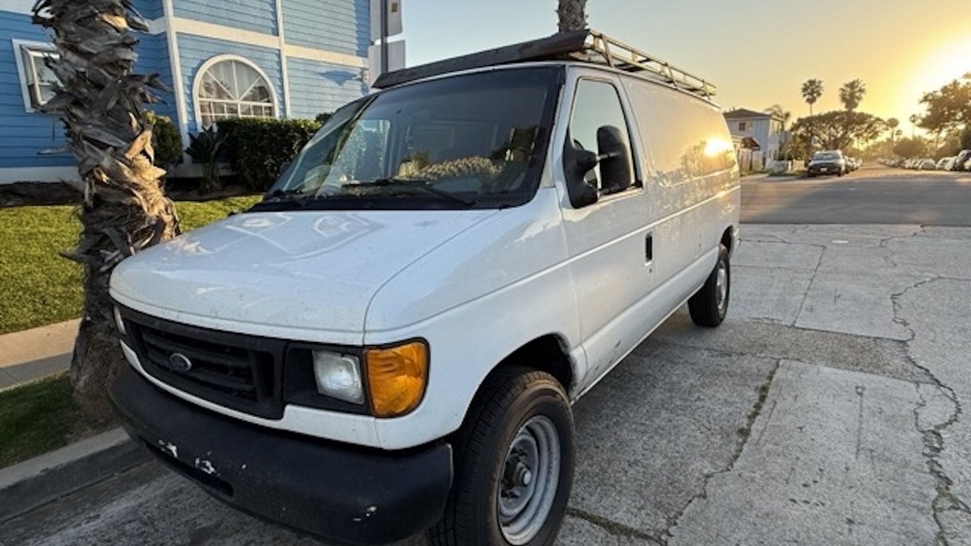 White cargo van with a roof rack parked on a sunlit residential street beside a blue house with an arched window; a palm tree stands nearby as the sunset casts warm light.
