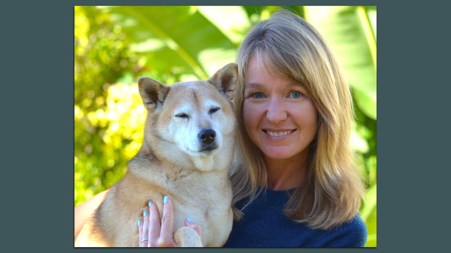 Smiling blonde woman in blue holding a calm tan and white Shiba Inu dog outdoors with green foliage blurred in the background.