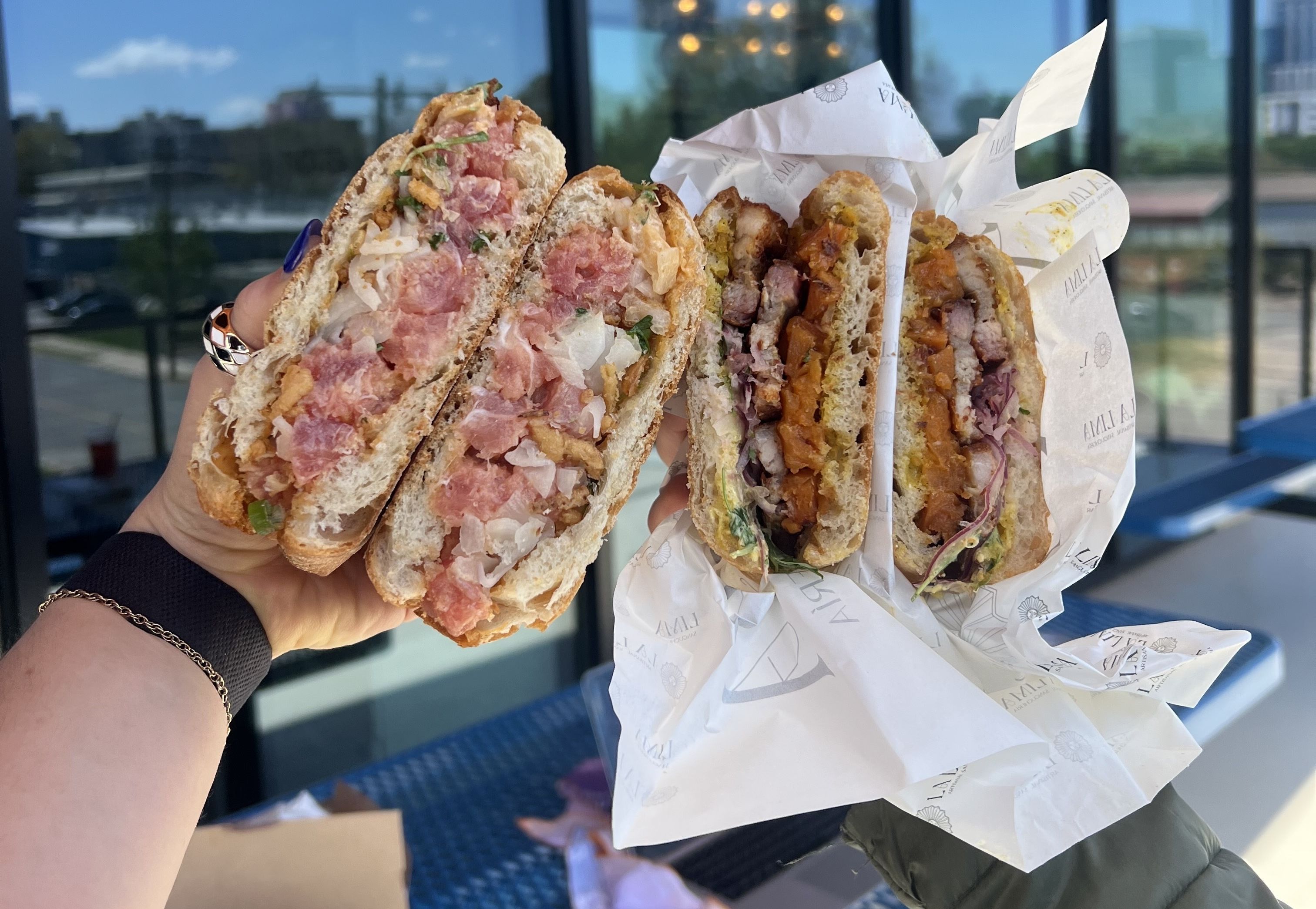 Two hands each holding a sandwich cut in half, revealing fillings like diced pink meat, onions, greens, and grilled meat with orange and purple vegetables inside crusty bread.