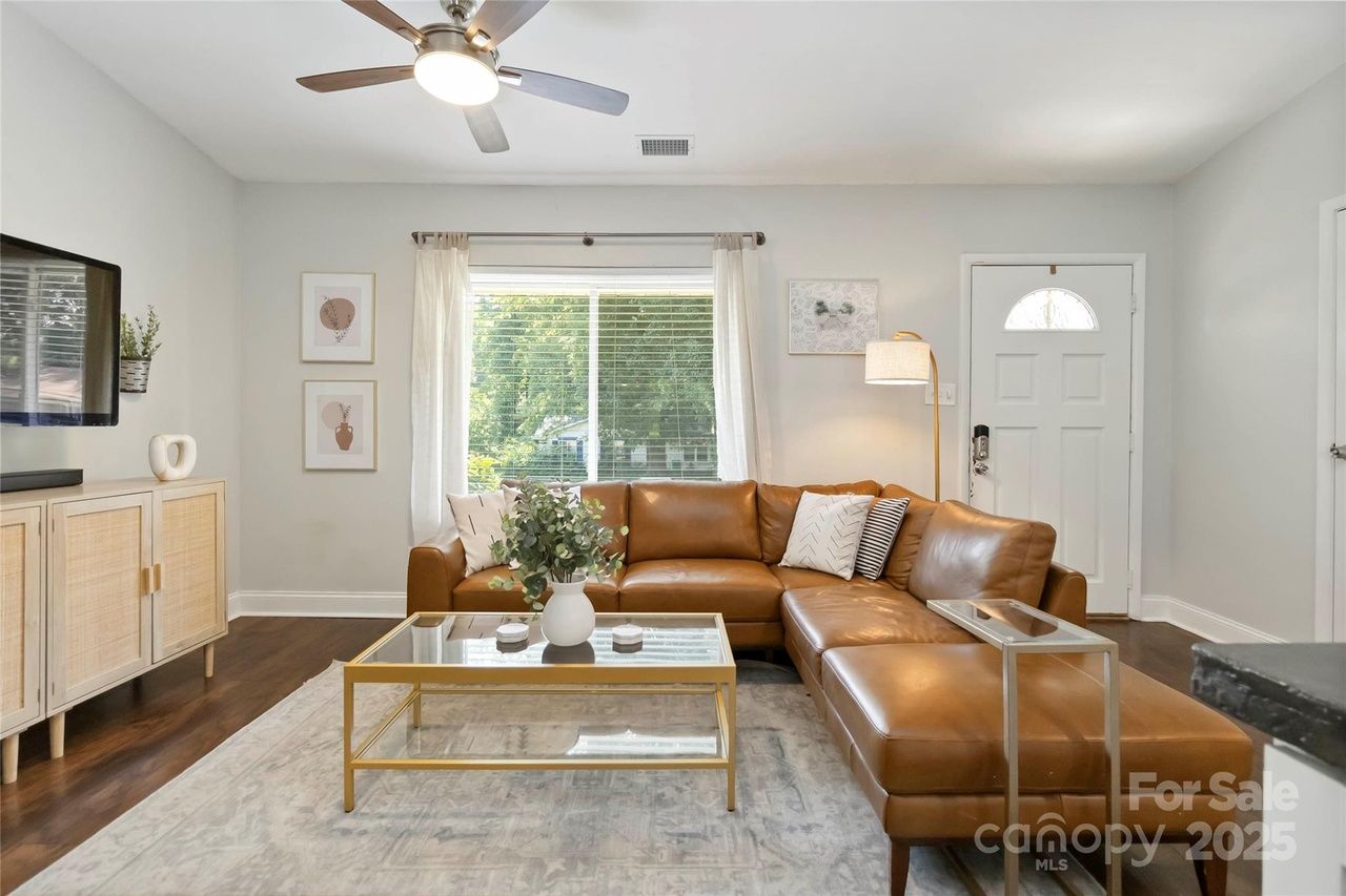 Bright living room with tan leather sectional sofa, glass coffee table with gold frame, large window with white curtains, ceiling fan, and wooden cabinet under wall-mounted TV.