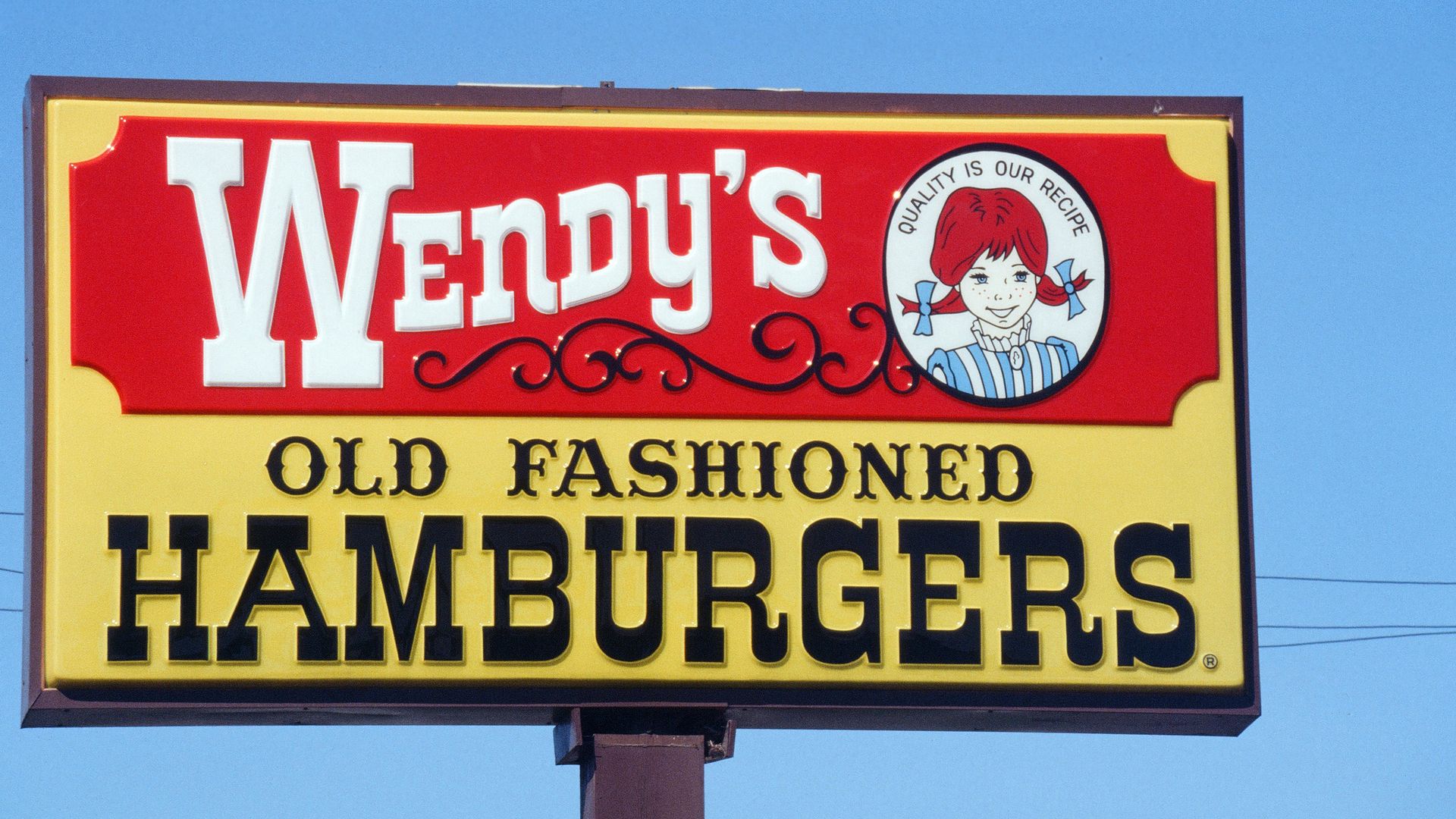 Sign for Wendy's fast food on a yellow board: red panel with large white 'Wendy's' logo, circular emblem of a girl, and bold black text 'OLD FASHIONED HAMBURGERS' beneath, against a blue sky.