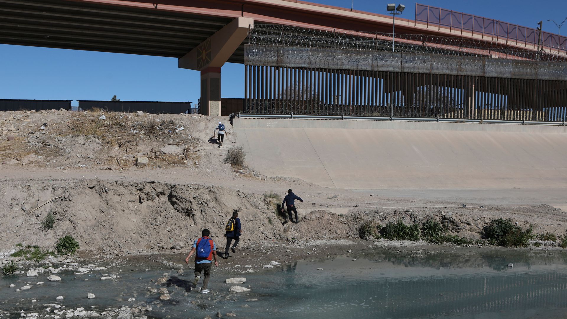 Migrants crossing water heading toward a bridge and border crossing