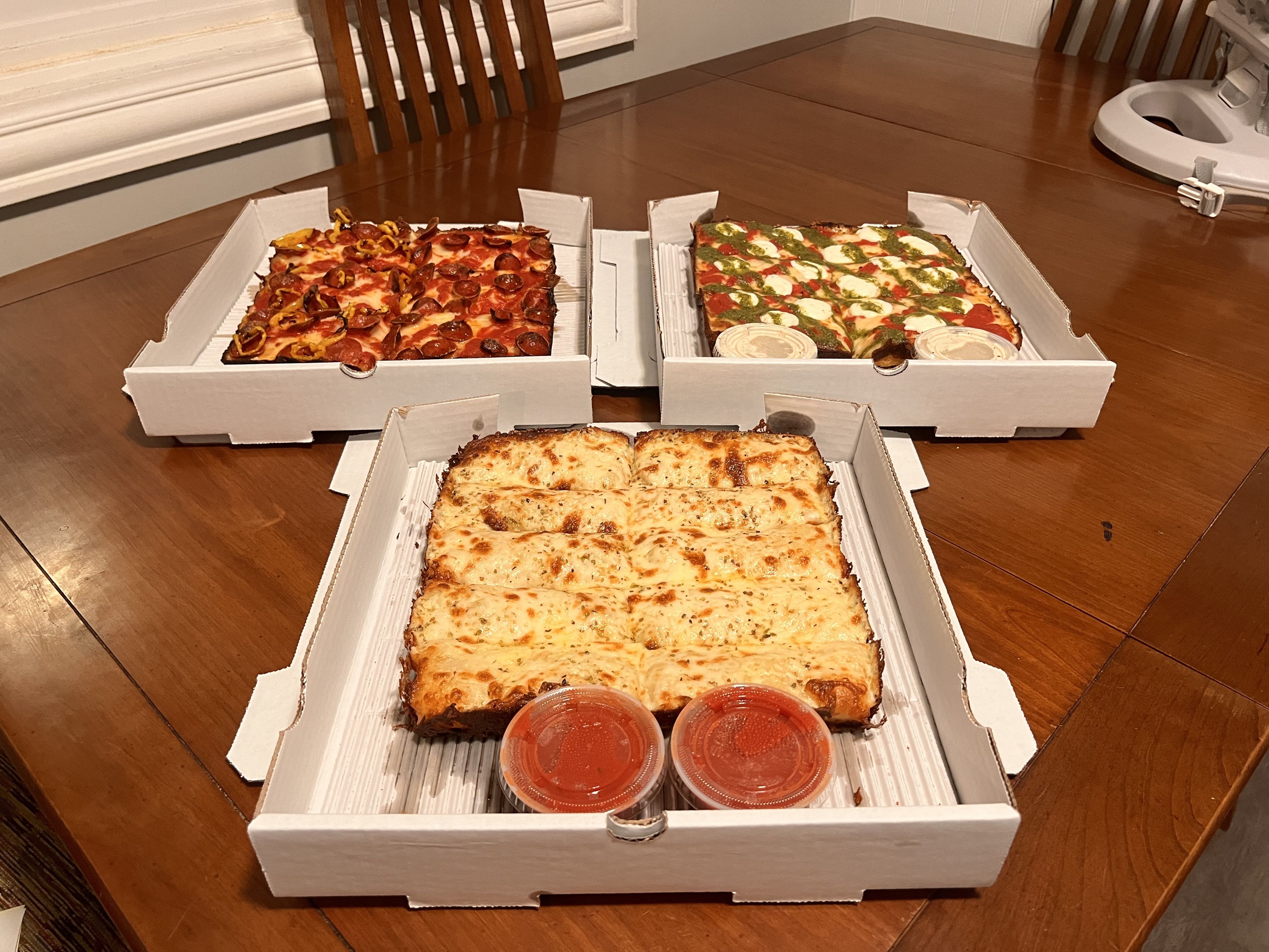 Three open pizza boxes on a wooden table with rectangular pizzas: cheese sticks with marinara sauce, pepperoni with yellow peppers, and mozzarella, pesto, and tomato.