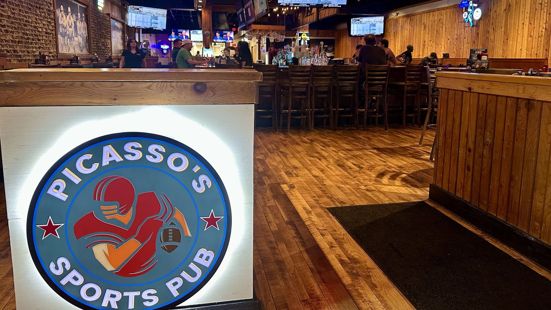 Rustic sports bar interior with multiple ceiling TVs showing games, wooden walls and bar with stools, a neon Stella Artois sign, and a large circular logo in the foreground.