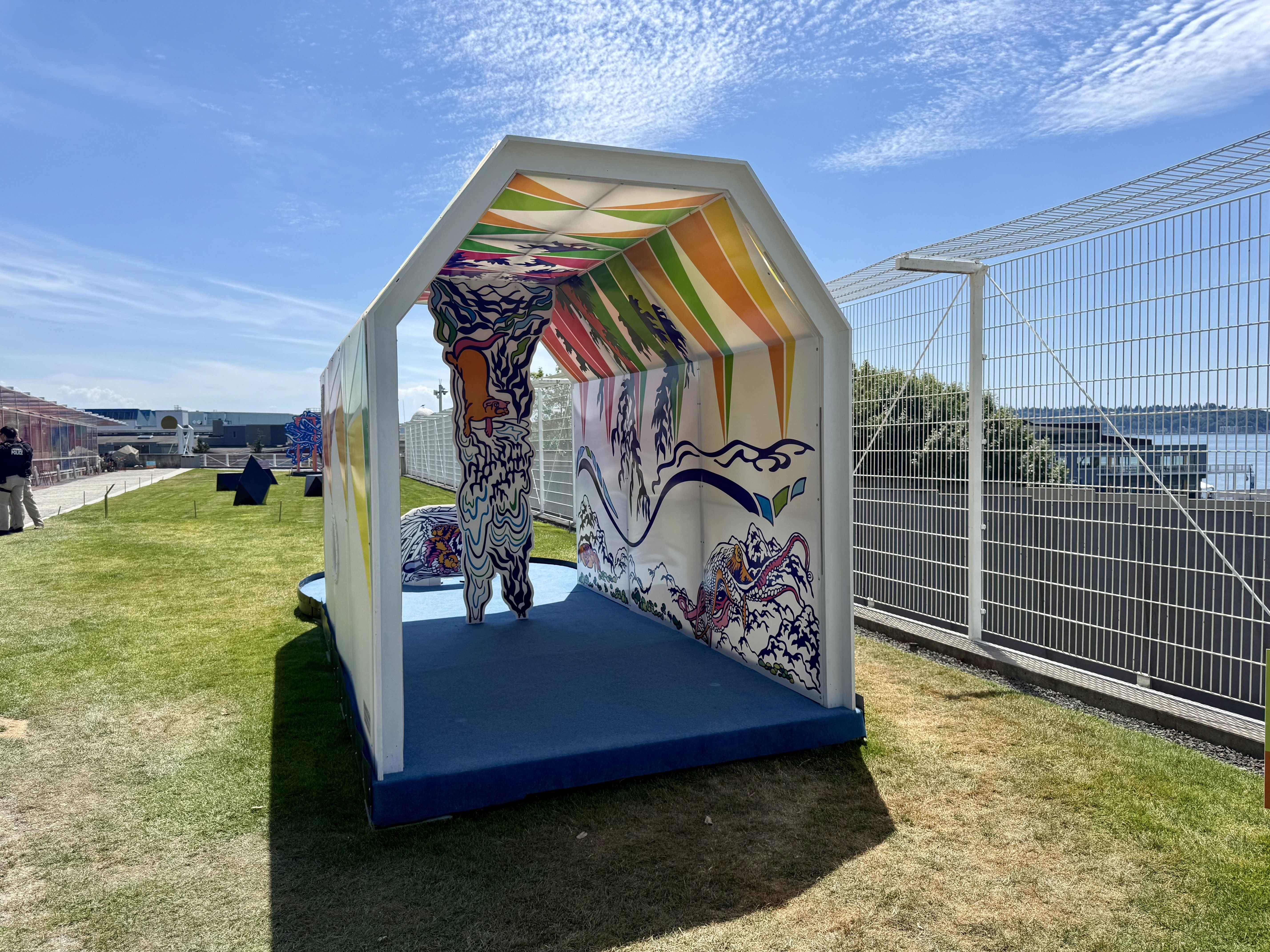 White pavilion with rainbow-striped interior and sea-art murals, set against a blue sky with wispy clouds. Green grass, a tall fence on the right, and a person in dark clothing on the left.