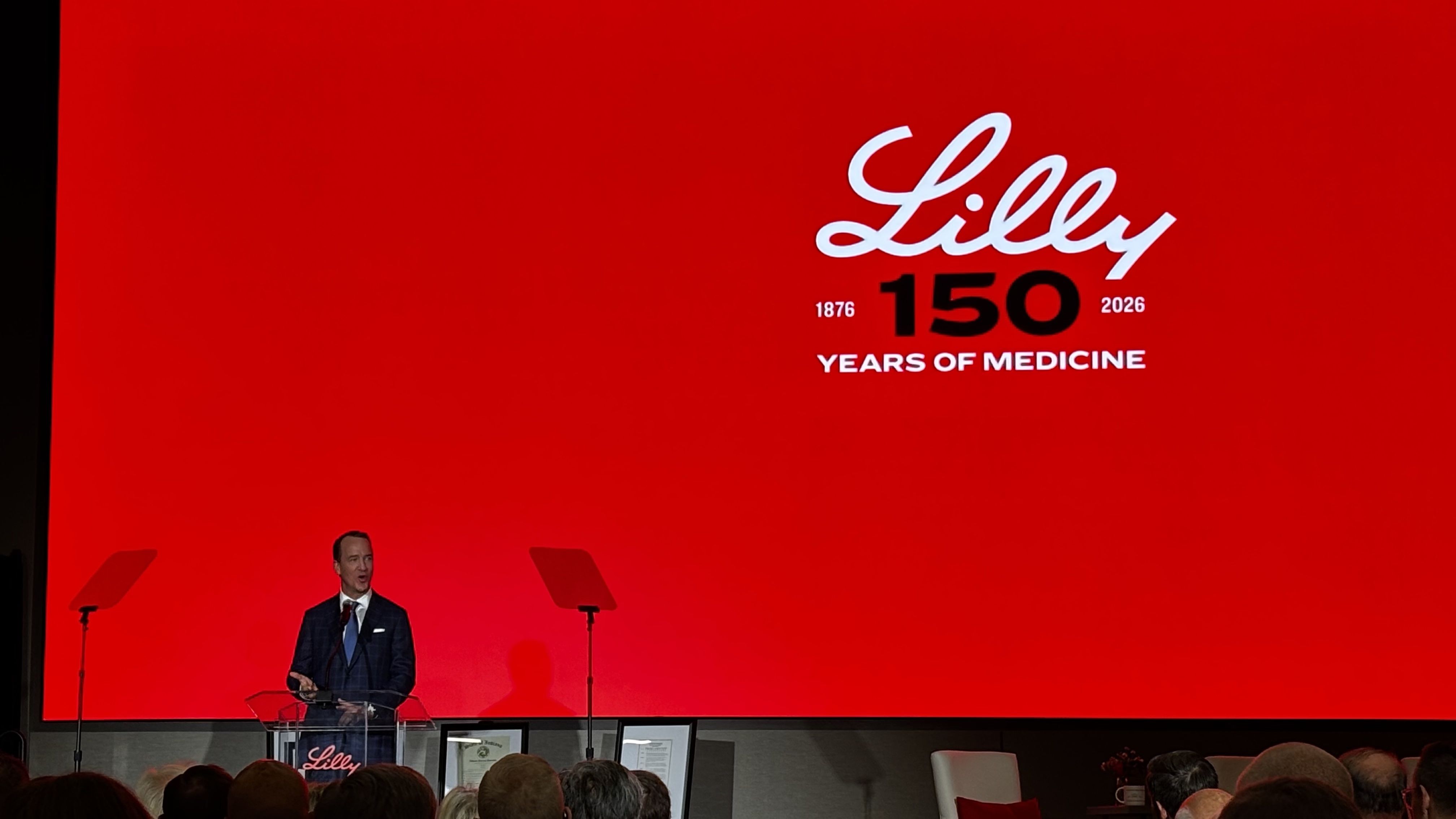 Speaker in a dark suit at a podium on a bright red stage; a large screen displays the Lilly logo, the number 150 with 1876–2026 and the title "YEARS OF MEDICINE." Audience silhouettes are in the foreground.