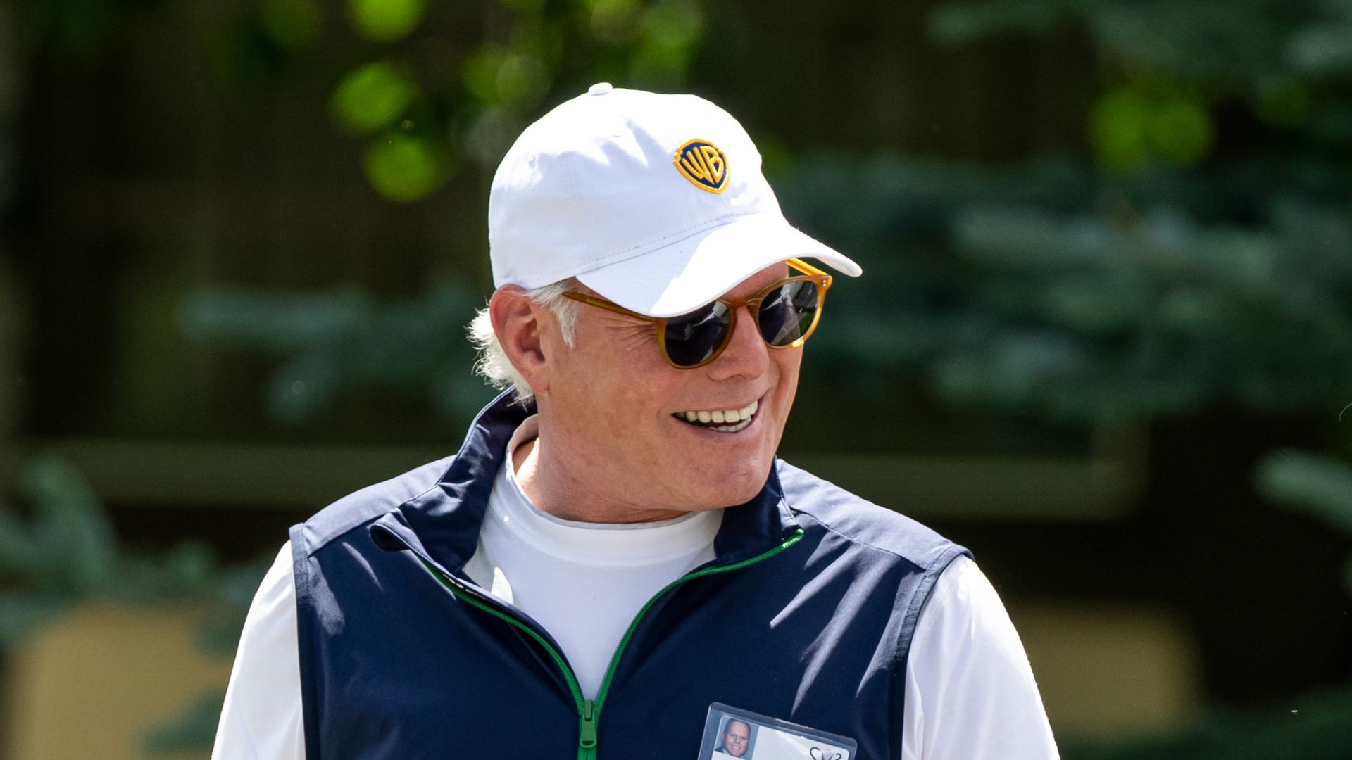 David Zaslav, president and CEO of Warner Bros. Discovery, wears a white Warner Bros. cap, sunglasses, and a vest, smiling and pointing toward the camera.