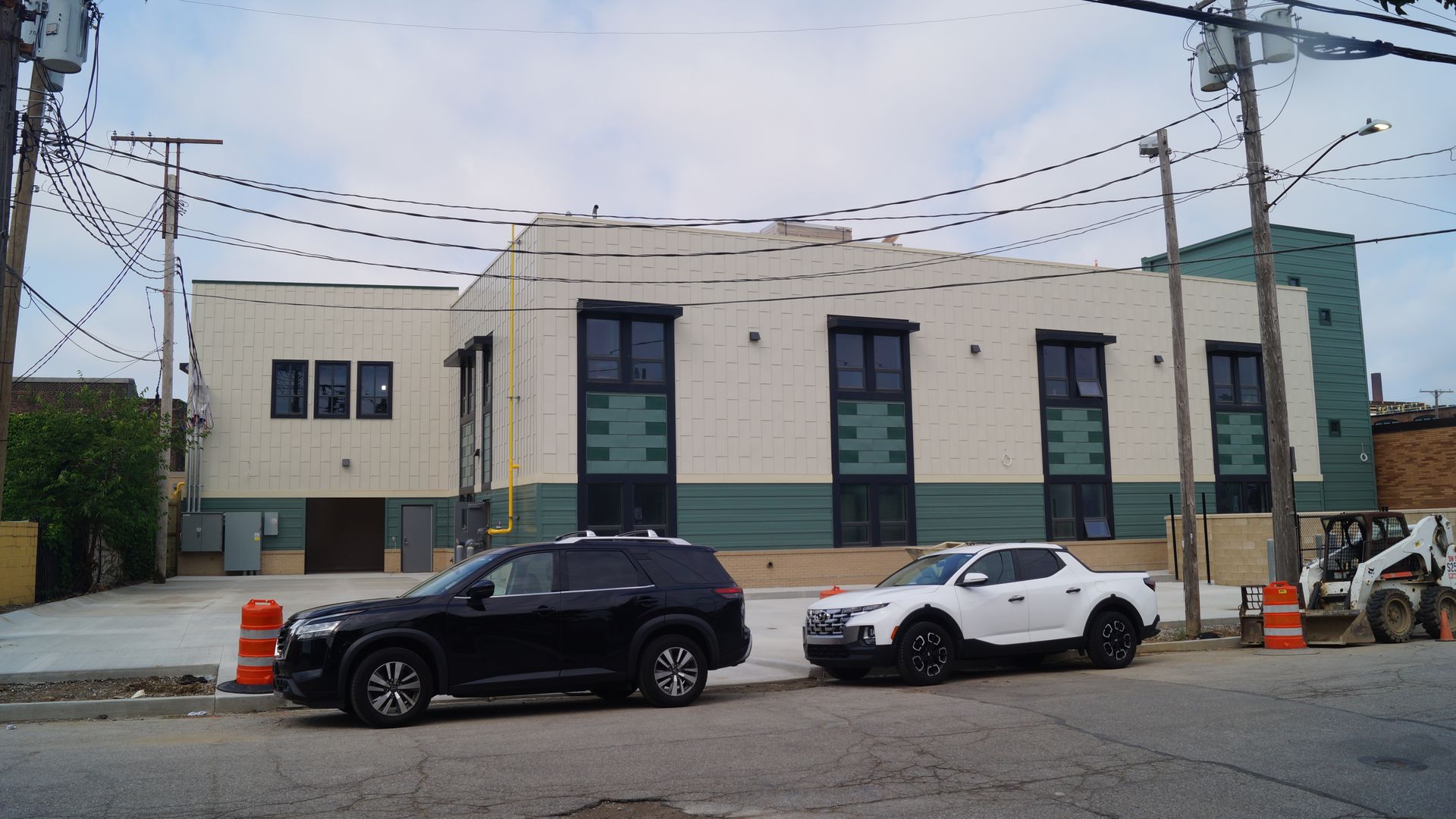 Beige two-story building with dark green base, numerous overhead power lines, and a construction site in front. Cars parked along the street; orange barriers and a small excavator visible on the right.