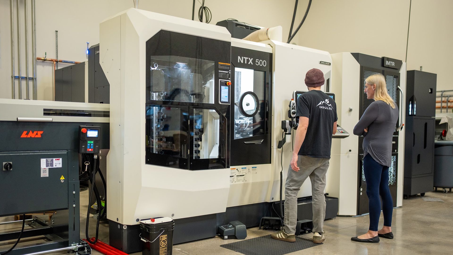 Two people stand at a large white CNC machine labeled "NTX 500" in a workshop. A man in a beanie and a woman in gray watch the control panel, with other equipment visible in the background.