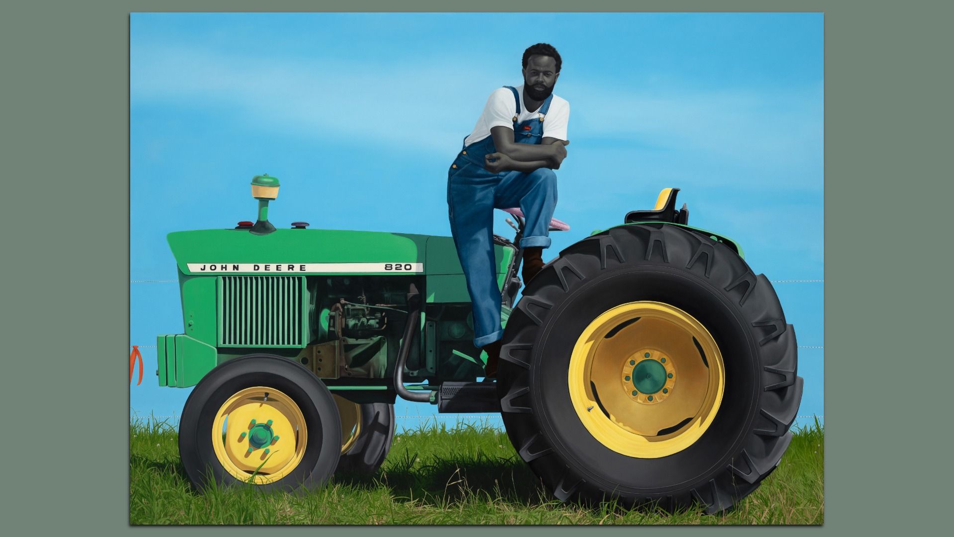 Bearded man in blue overalls and a white shirt sits atop a large green John Deere tractor with yellow rims, against a bright blue sky and grassy field.