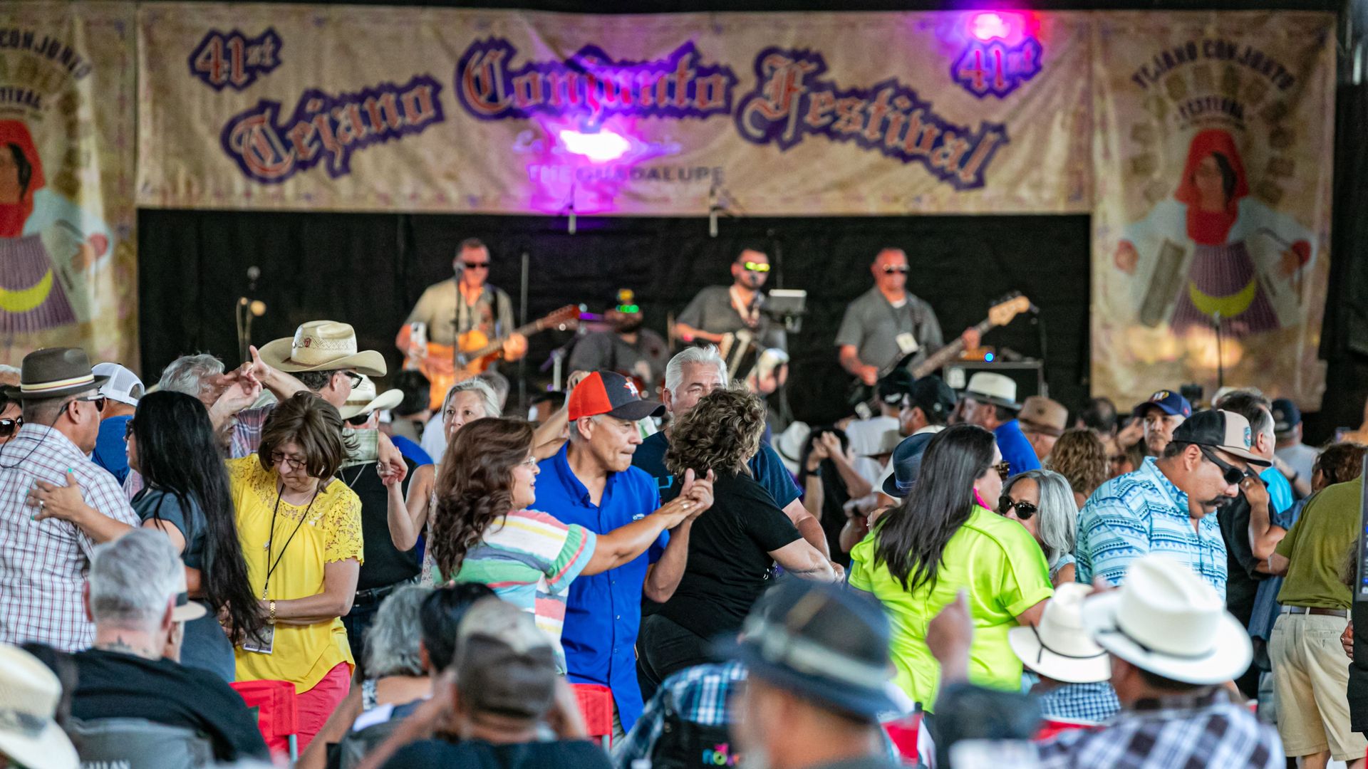 People dancing together in front of a stage with musicians playing and a sign that reads "41st Tejano Conjunto Festival" in the background.