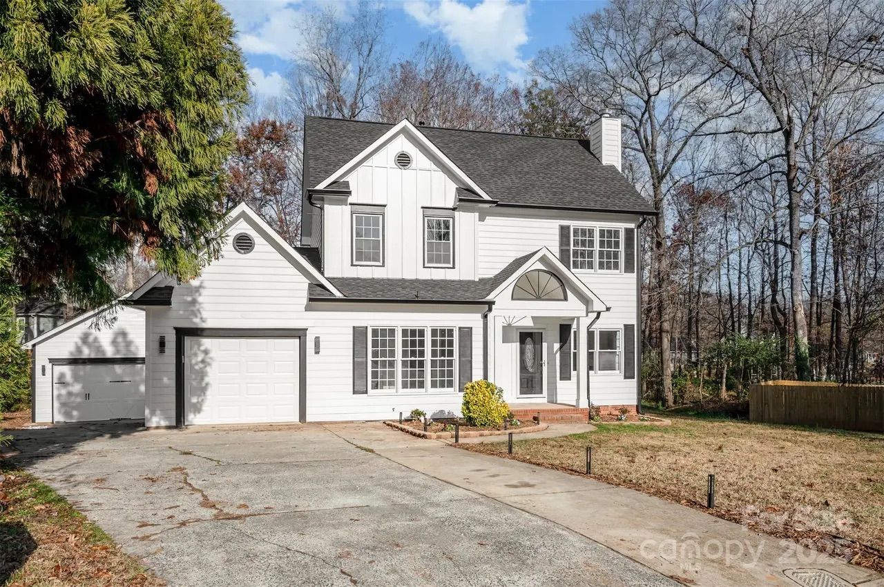Two-story white house with black shutters and roof, attached garage, concrete driveway, and bare trees in the background under a partly cloudy blue sky.