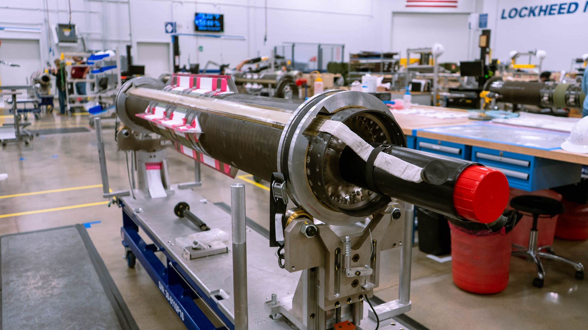 Long cylindrical mechanical device with a black tube and red cap on a metal stand in a spacious, well-lit workshop with tool benches and equipment in the background.