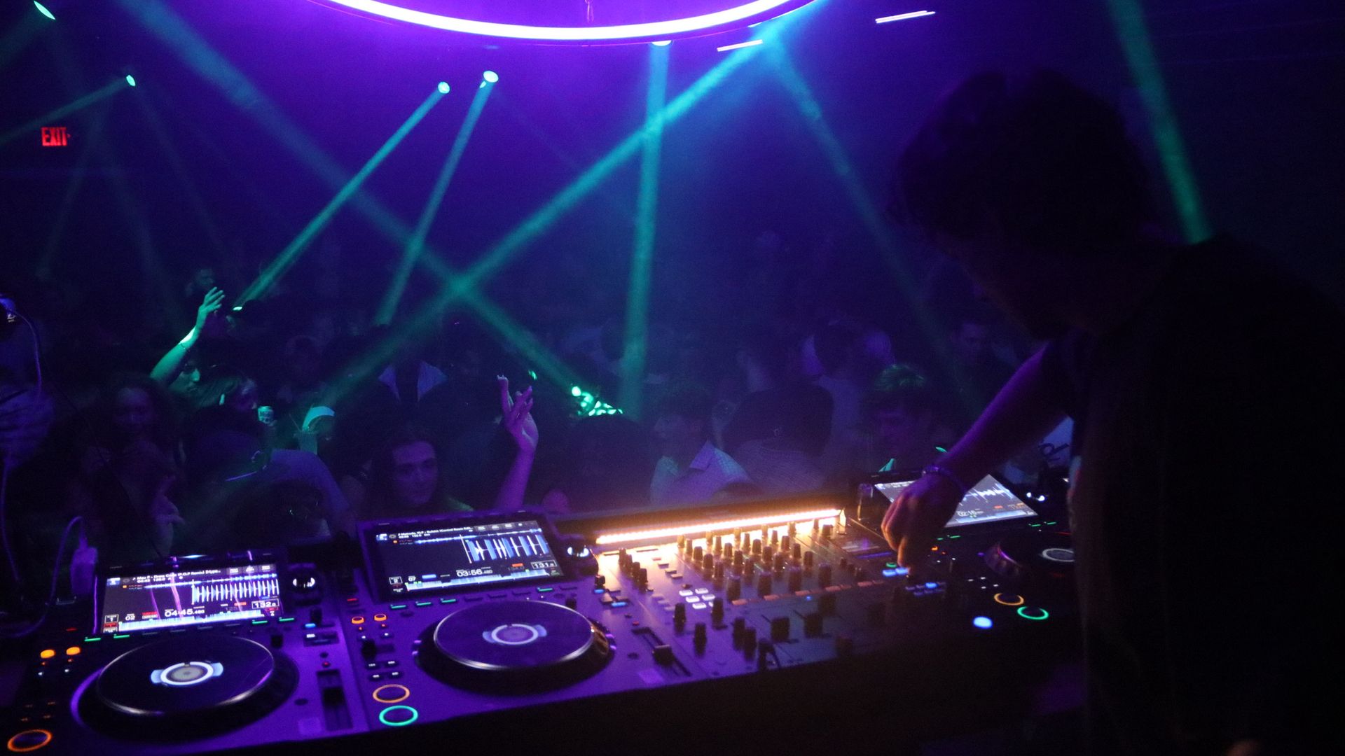DJ booth in the foreground with turntables and mixer; purple and blue club lighting, green laser beams across the crowd, a dark dance floor behind, and a red EXIT sign on the left.