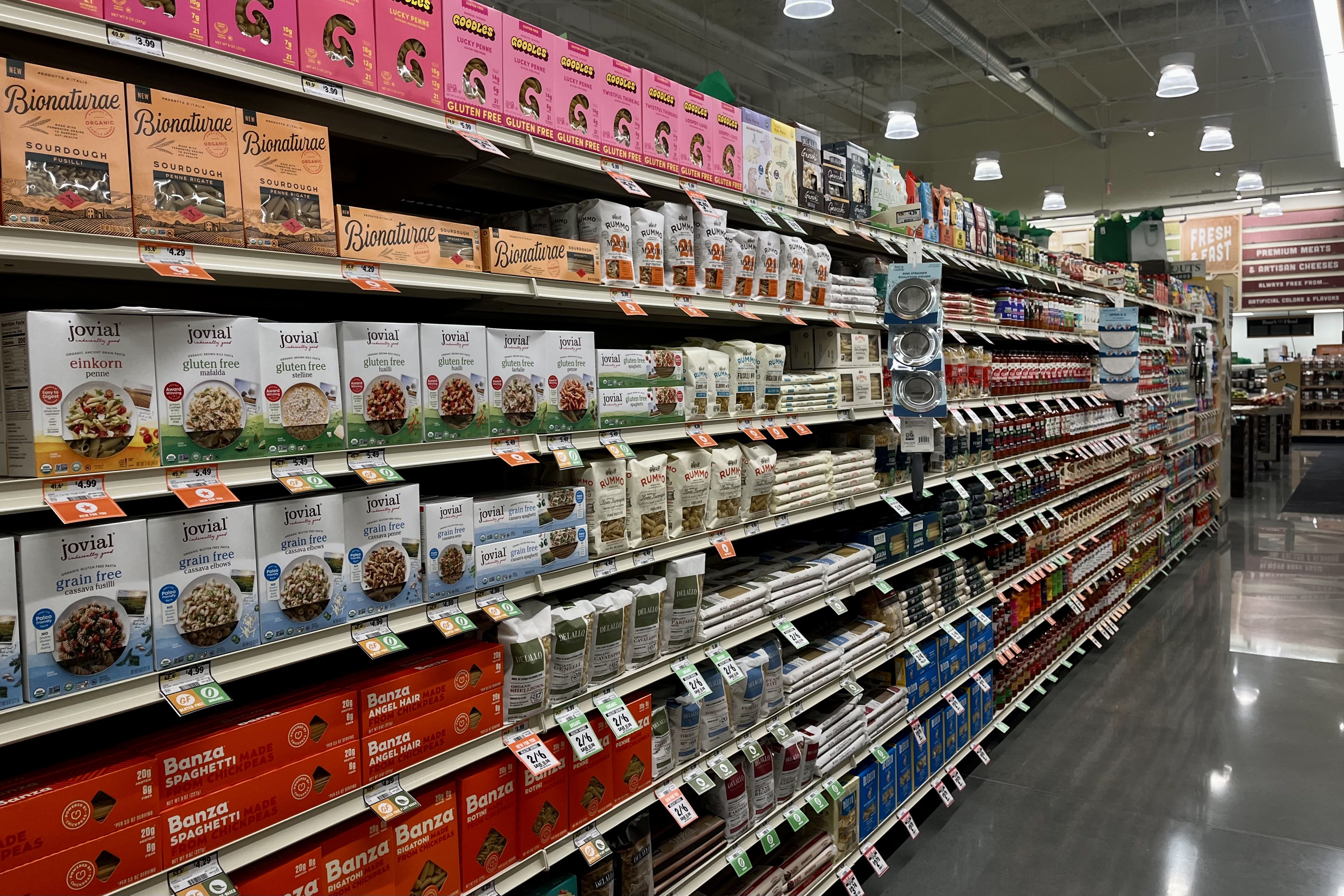 A grocery store aisle with shelves stocked with various pasta brands in colorful packaging, including gluten-free and organic options, under bright ceiling lights on a shiny floor.