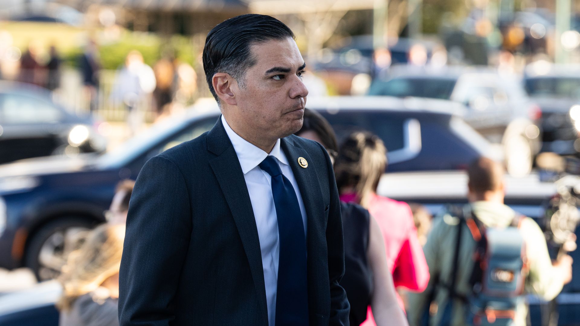 Rep. Robert Garcia, wearing a blue suit and standing outside in front of cars and people.