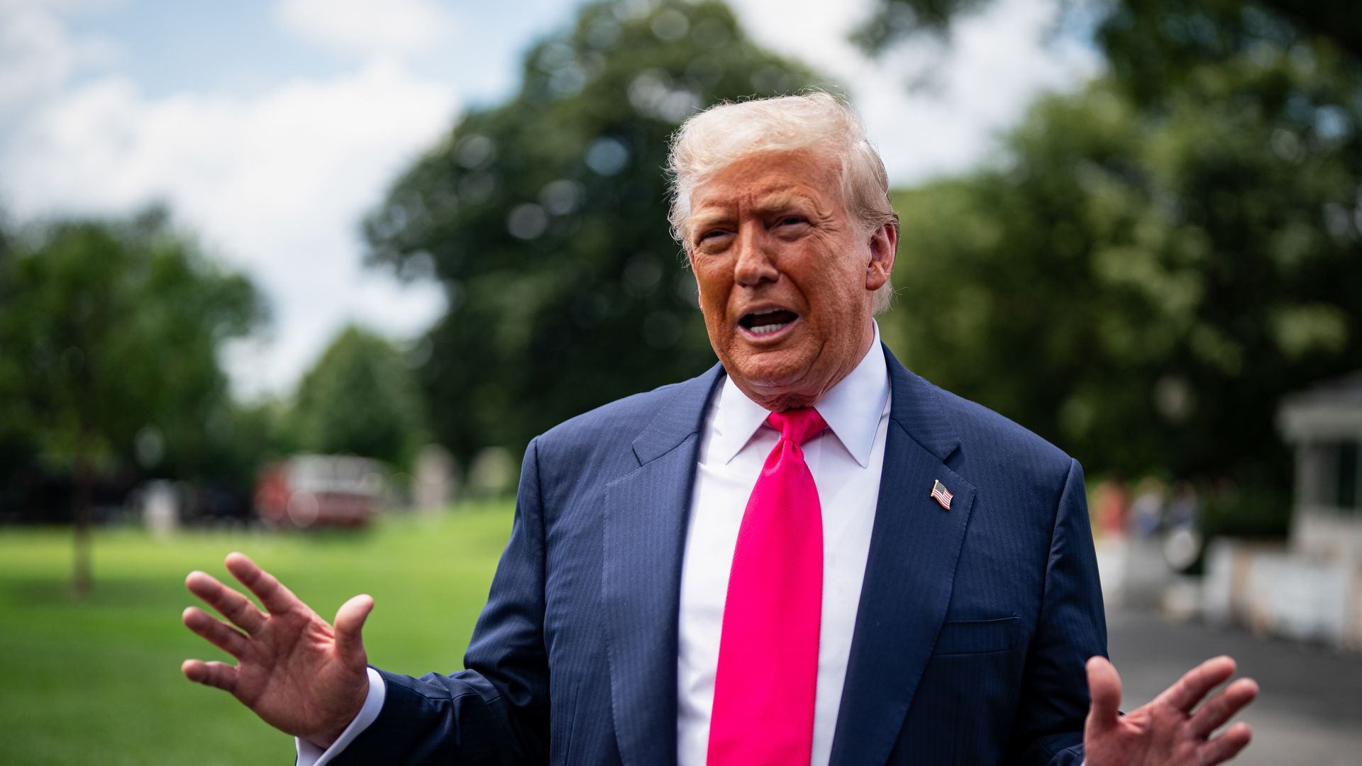 Donald Trump in a dark blue suit, bright pink tie, and white shirt speaking outdoors with hands raised, green grass and trees in background under partly cloudy sky.