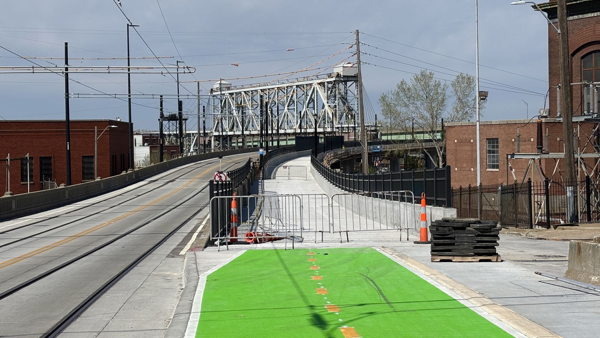 Urban street with rail tracks on the left, a steel bridge in the distance, orange cones and metal barriers, and a bright green bike lane in the foreground beside brick buildings.