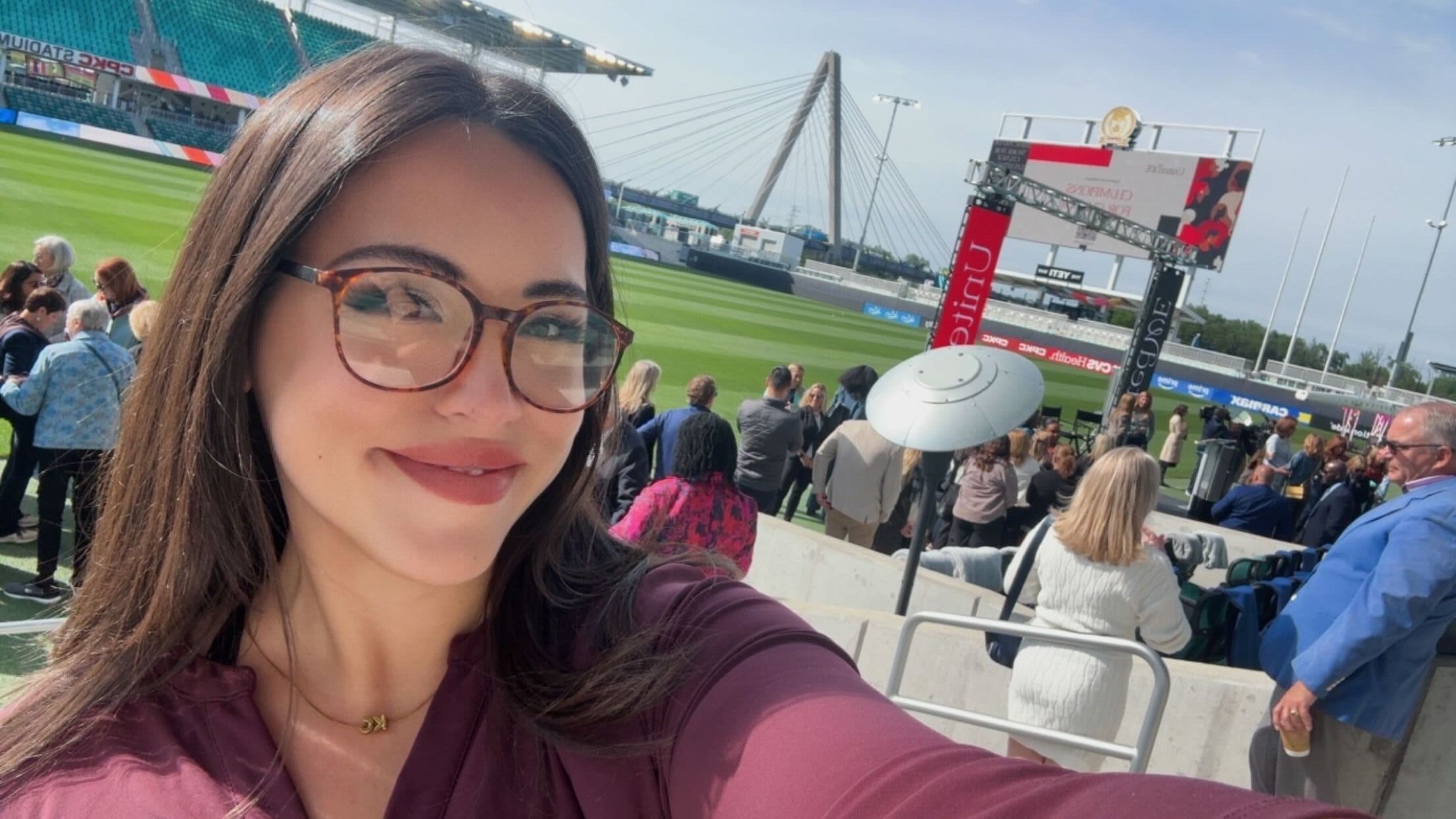 Reporter Abbey in tortoiseshell glasses in the foreground at a sunny stadium. Behind her, a green field, seated crowd, a large screen, and a cable‑stayed bridge.