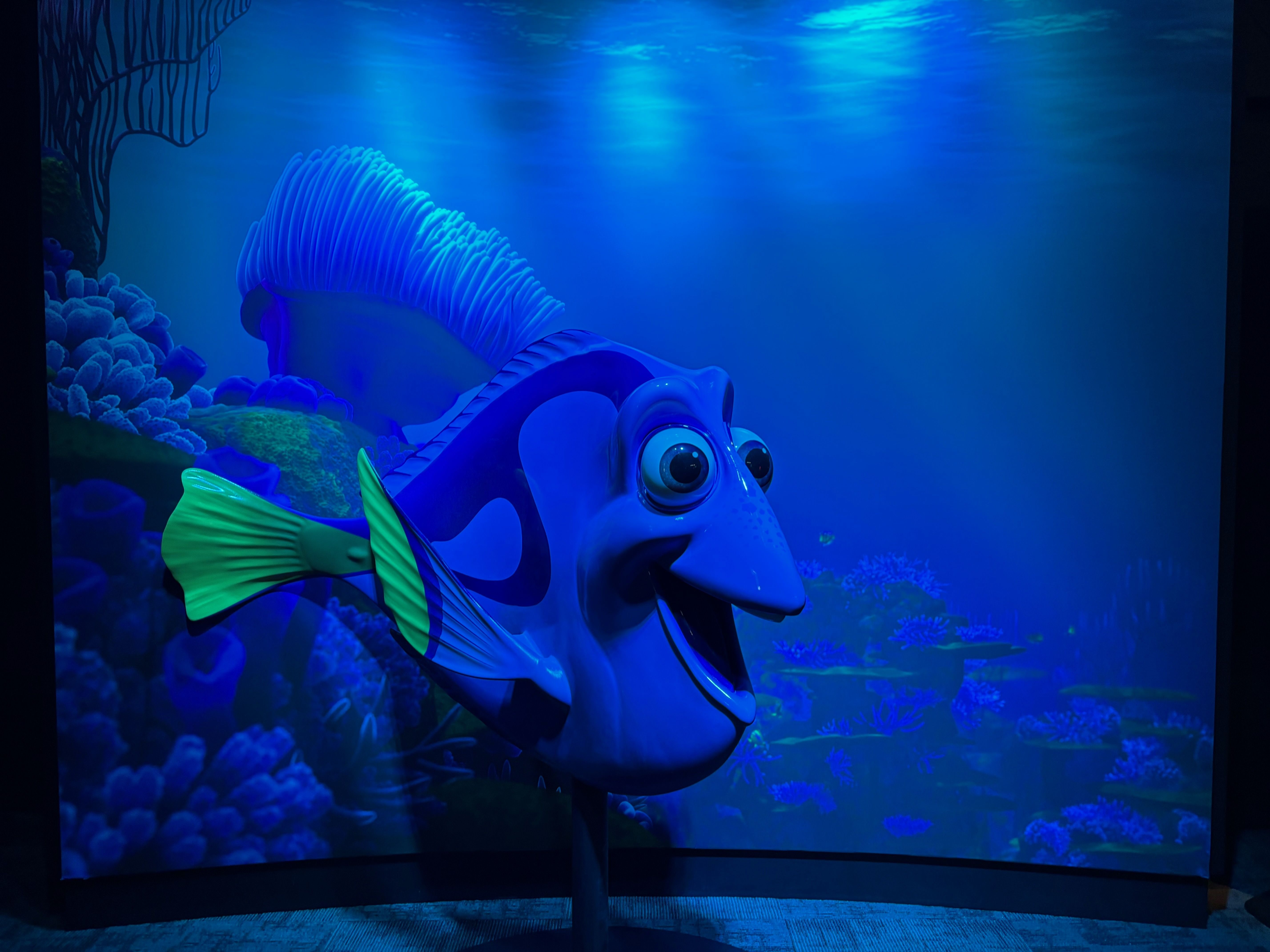Blue and yellow animated fish character with large eyes and open mouth, set against a deep blue underwater coral background with glowing light effects.