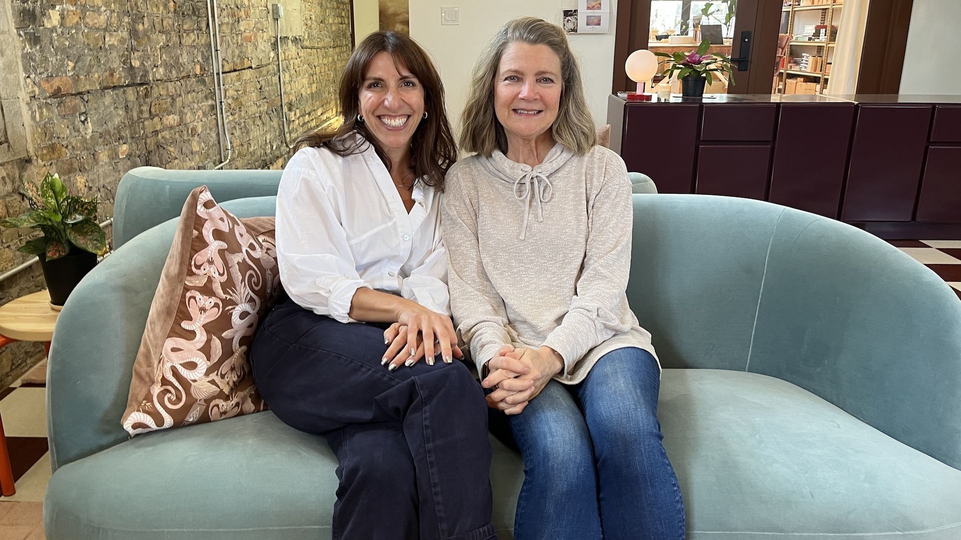 Two smiling women sit close on a teal curved sofa in a modern lobby; one wears a white shirt and dark pants, the other a light gray hoodie, with a stone wall, plant, and maroon cabinets behind them.