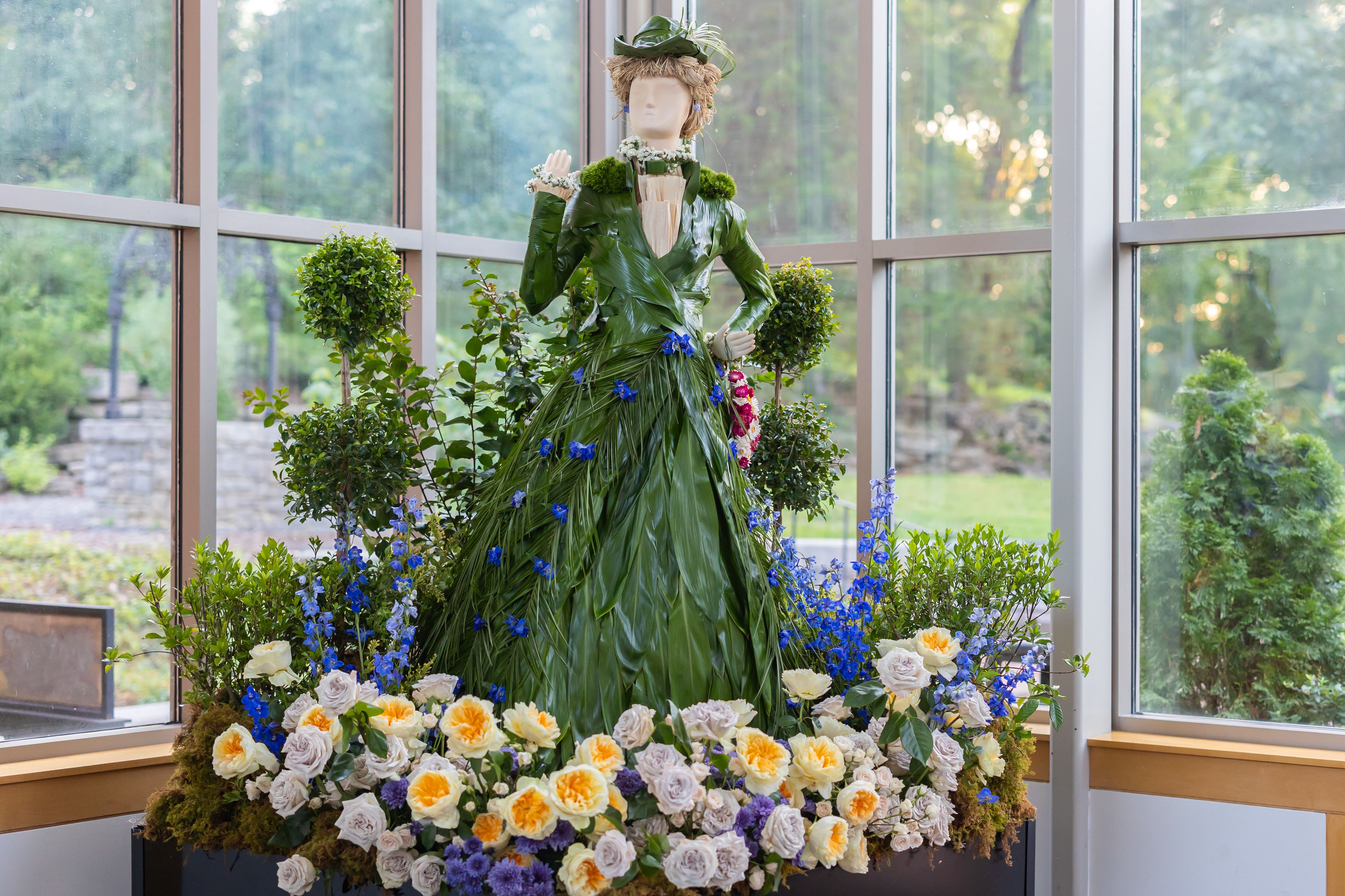 A life-size mannequin in a gown of green leaves, with a leaf hat and floral collar, posed among potted shrubs and a bed of yellow and white roses inside a glass conservatory.