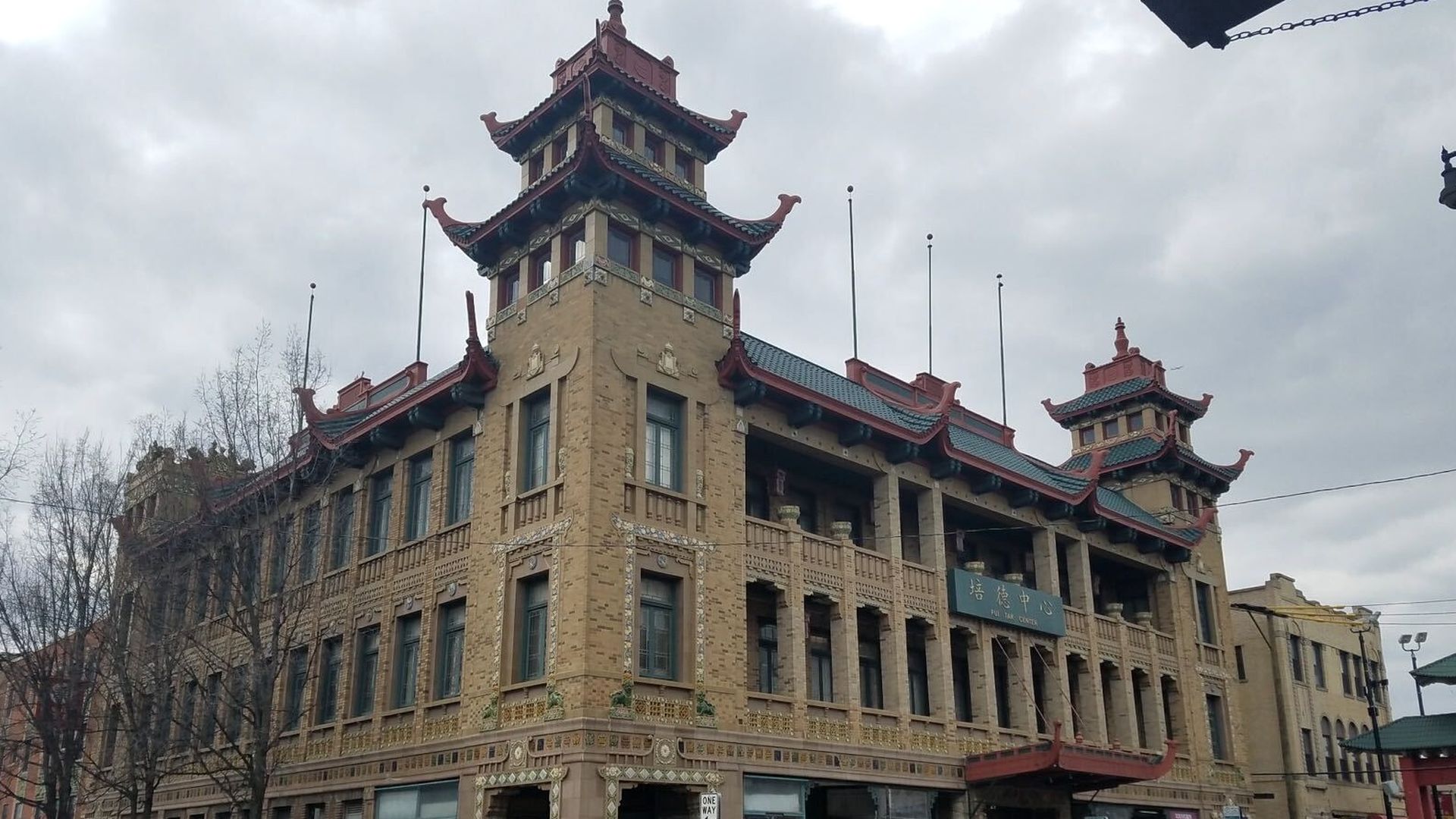 Beige brick building with two pagoda-style towers, red curved roofs, and decorative trim. Large windows line the facade; cars parked along the street under a cloudy sky.